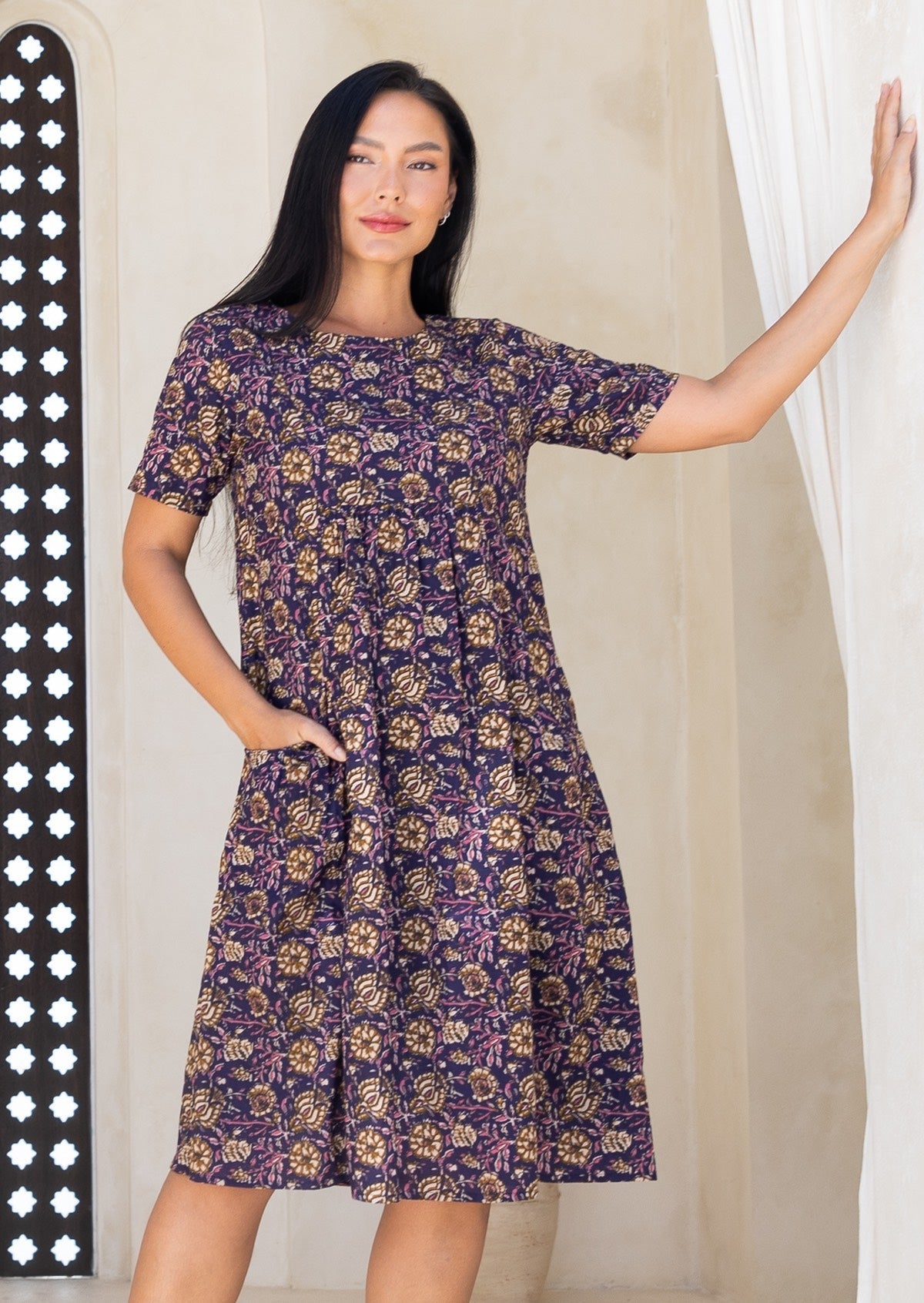 Woman wearing a purple floral dress with hand in pocket, against a neutral background