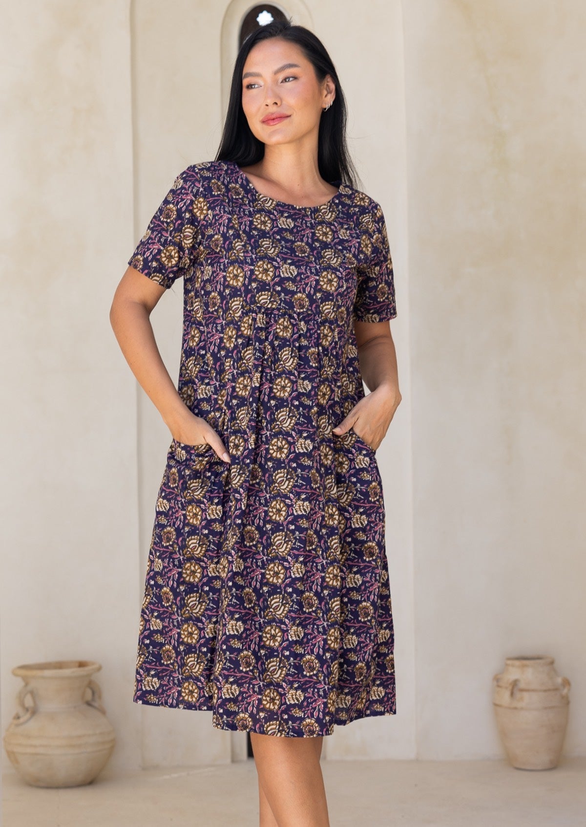 Woman wearing a purple floral dress with hands iin pockets, standing against a plain wall.