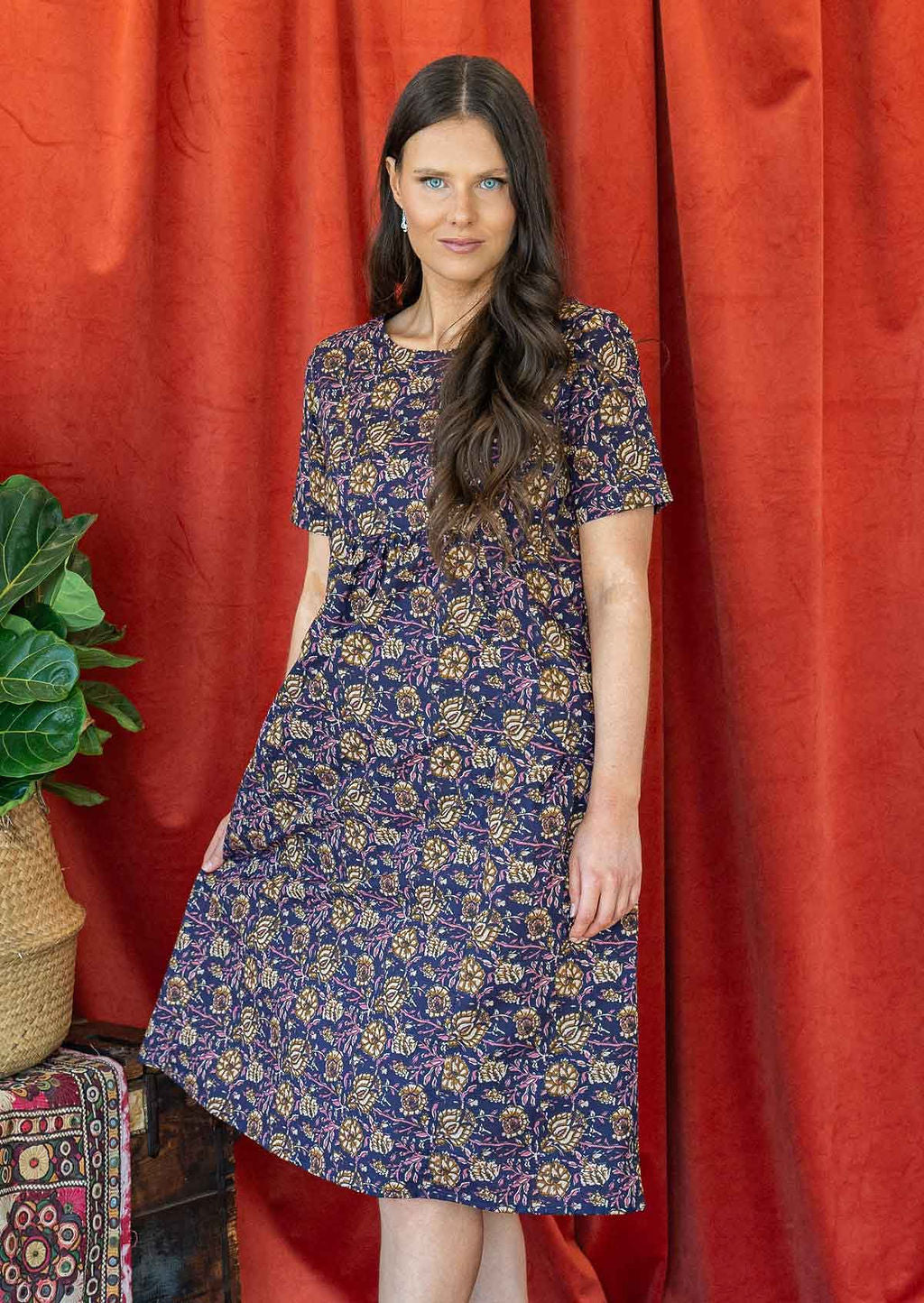 Woman wearing dark purple based floral printed cotton dress with an empire waistline and deep pockets.