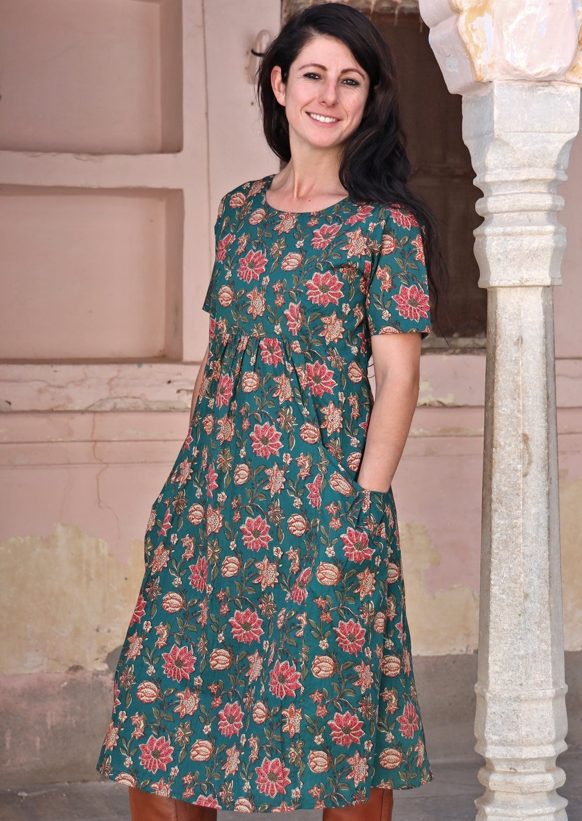 Model wears deep green floral printed cotton dress with simple round neckline and hands in the pocket.