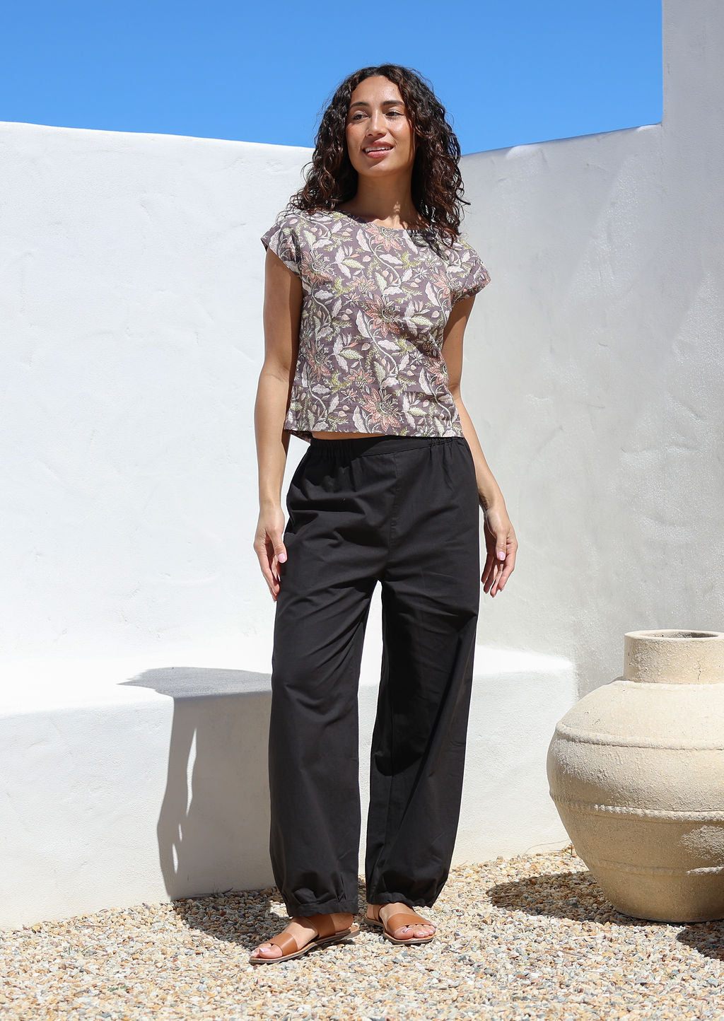 Woman standing in sun wears a floral cotton top with wide leg black cotton pants with elastic either side of waist and pleated ankles