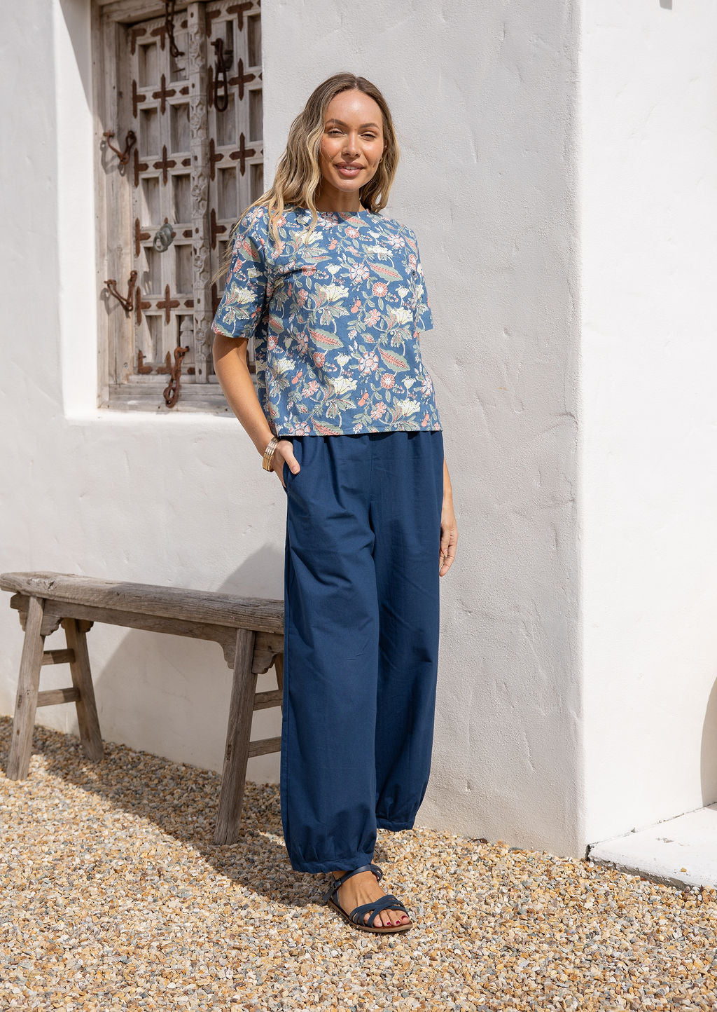 Woman stands wearing a dark blue 100% cotton wide leg harem pants with pleats at the ankles, elastic side waist and hand in pocket.