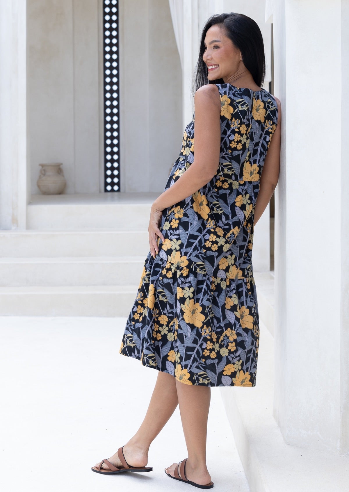 Woman wearing a black based midi length floral cotton dress with button back closure; standing in a bright, minimalistic setting.