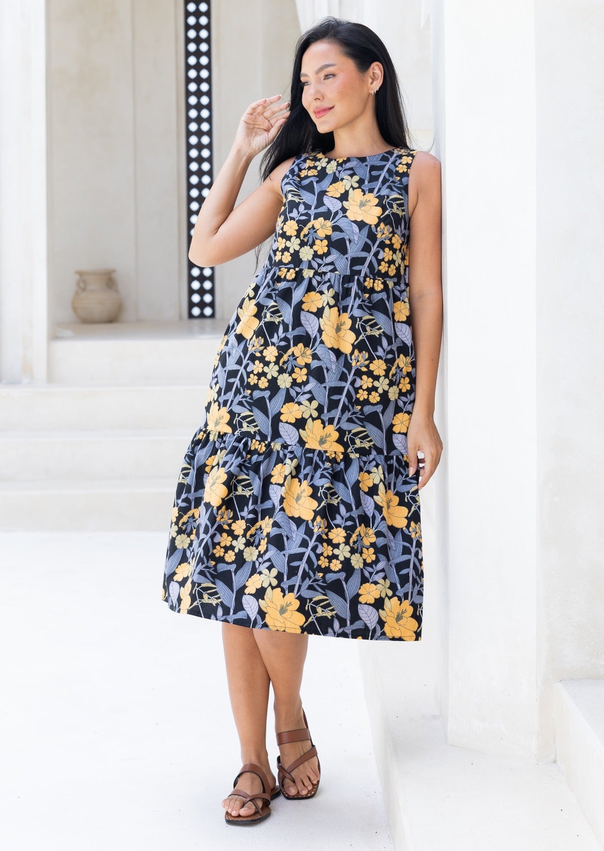 Model poses in sleeveless, black based floral print cotton midi dress with tiered design, pockets and high round neckline.
