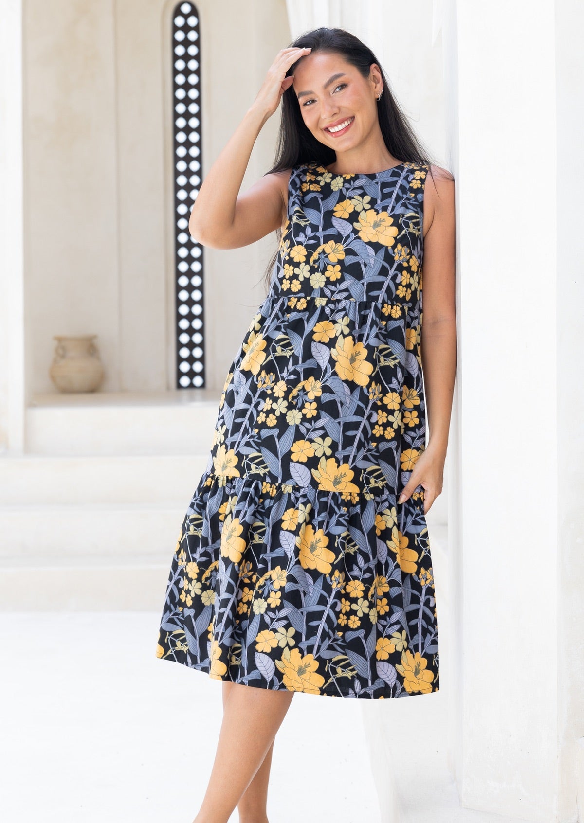 Model wears 100% cotton sleeveless midi dress with high round neckline, tiered design and pockets in black based yellow and grey floral print