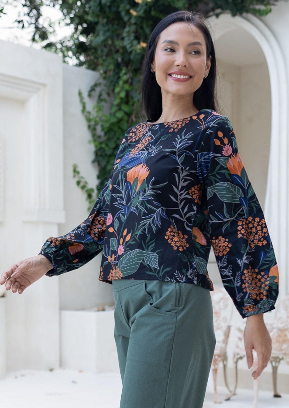 Woman wearing a dark floral, loose-fitting, long sleeved cotton top with a wide round neckline in Queen Protea print.