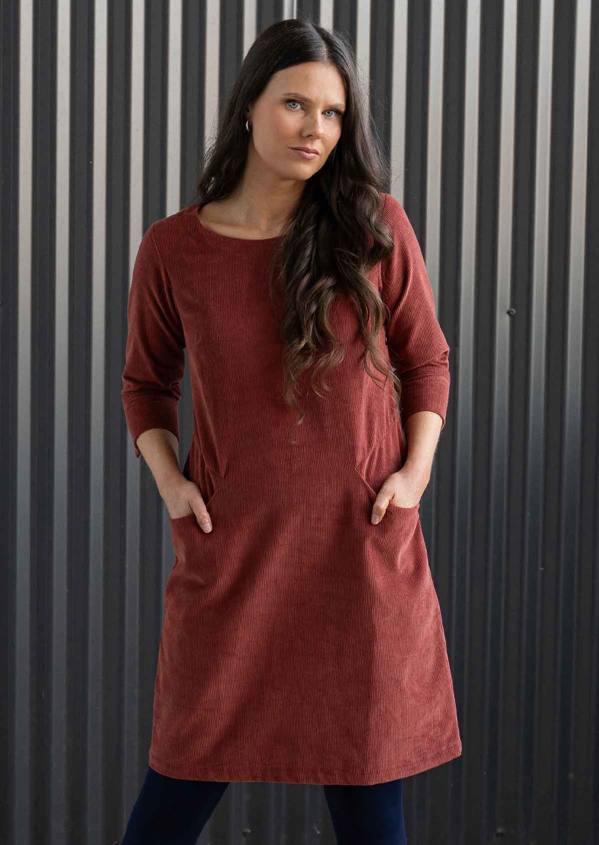 Woman wearing rust coloured cotton corduroy dress with sleeves over navy blue leggings and boots with hands in pockets