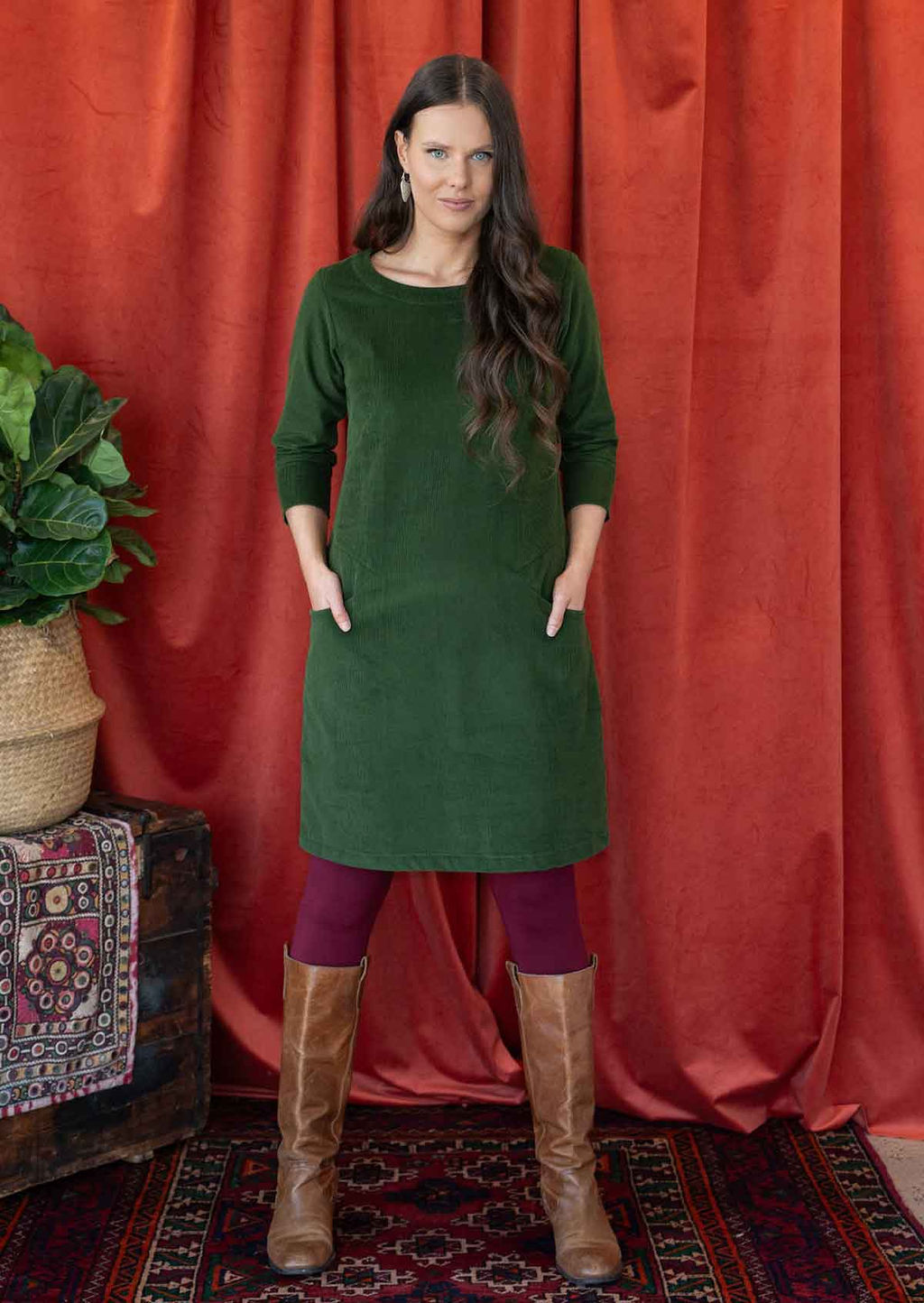 Woman wearing green corduroy A-line dress with 3/4 sleeves with hands in pockets over leggings and boots