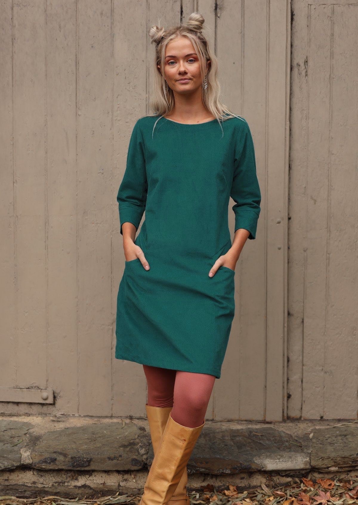 Woman with blond hair wearing Teal corduroy dress over pink leggings with 3/4 sleeves with hands in pockets