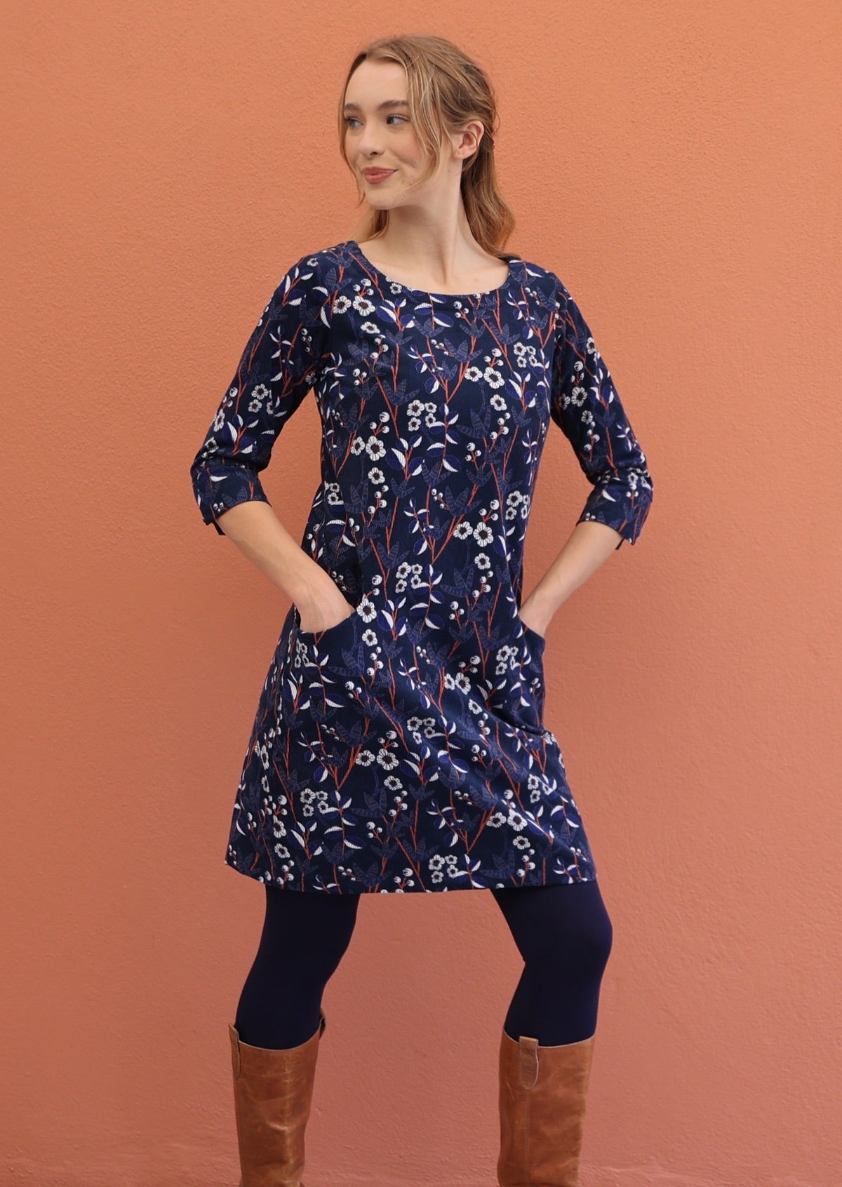 Woman wears round neck, 3/4 sleeve corduroy dress with hands in pockets, paired with navy blue leggings and boots