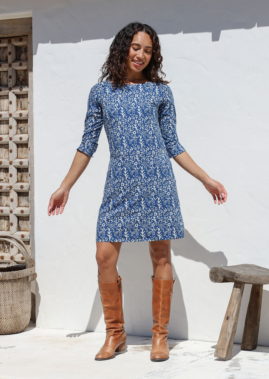 Woman wearing a blue cotton floral dress and brown boots standing against a white wall.