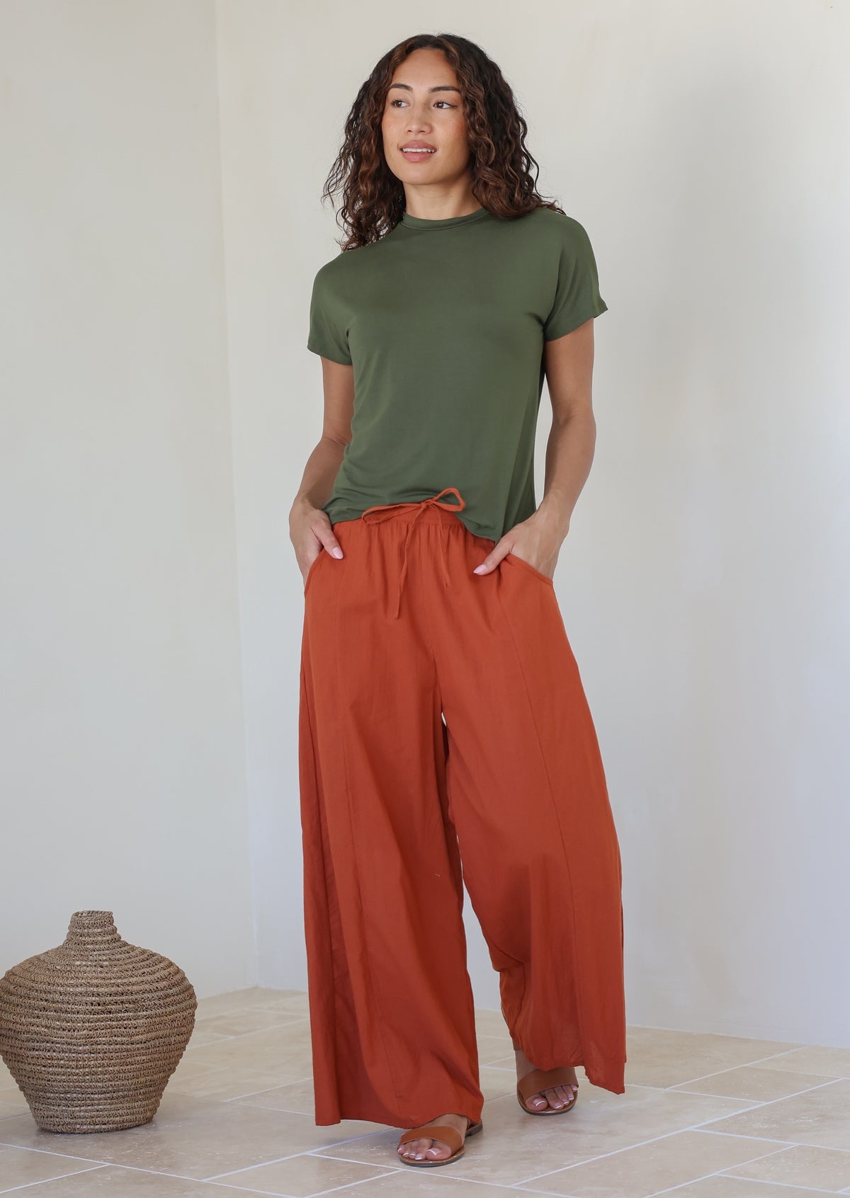 Woman stands in front of white wall wearing lightweight wide leg cotton pants in brick orange shade with pockets