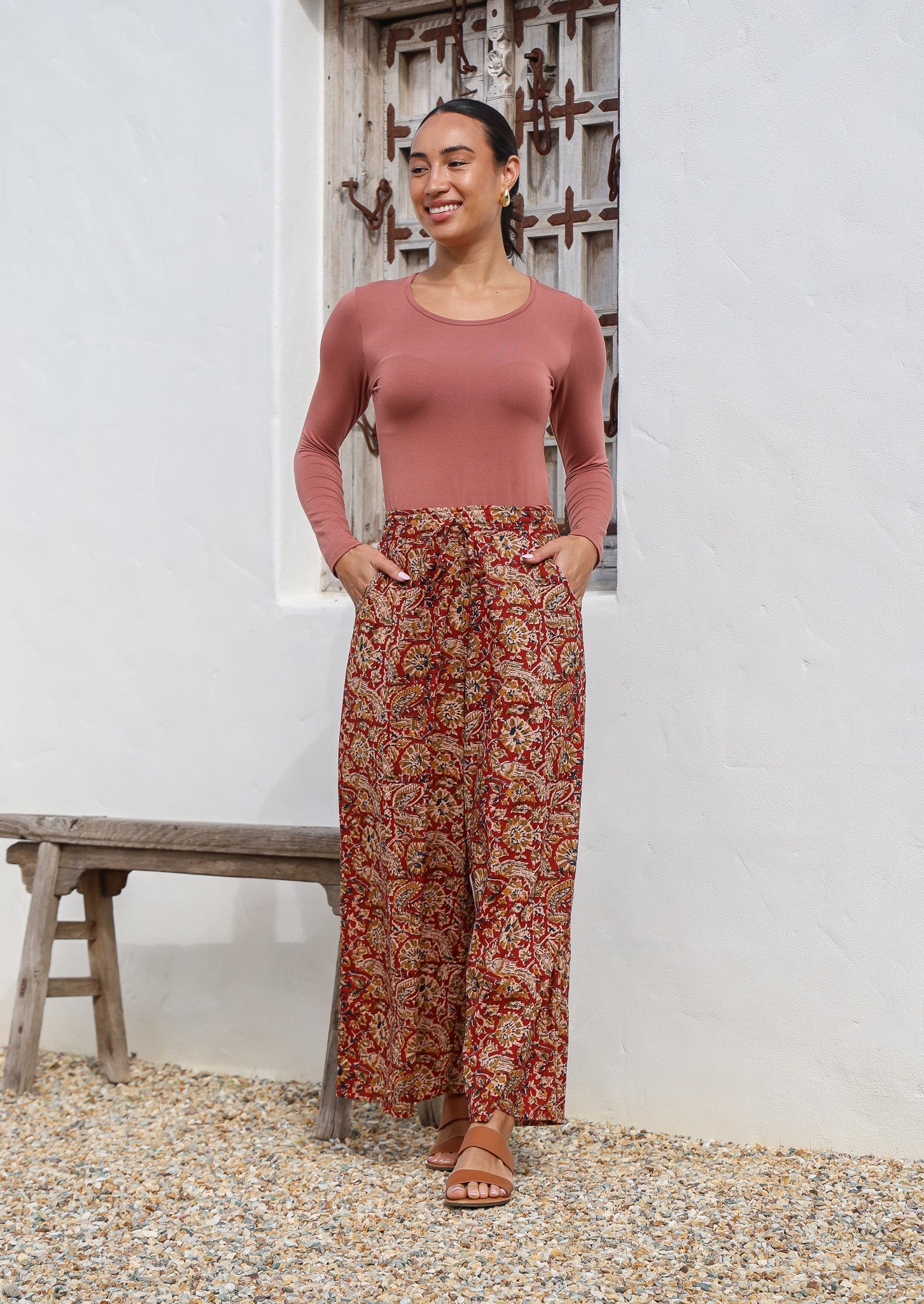 Model poses with hands in pockets of wide leg cotton pants with delicate floral print in earthy tones and red base.