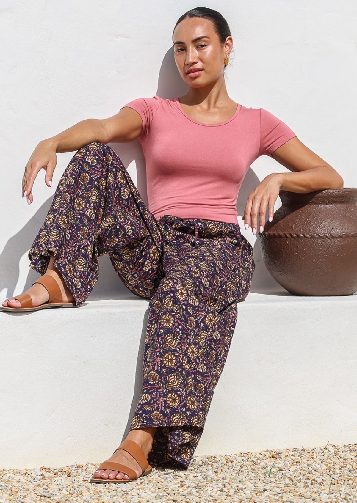 Model sitting in a relaxed pose wearing 100% cotton wide leg pants with pink, cream and light brown florals on dark purple base.