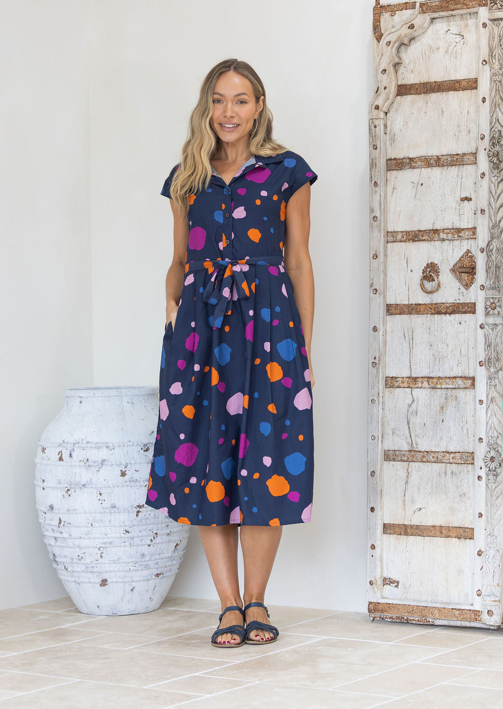 Woman wearing a cotton buttoned bodice with collar, navy dress with multi-coloured spots with hand in pocket