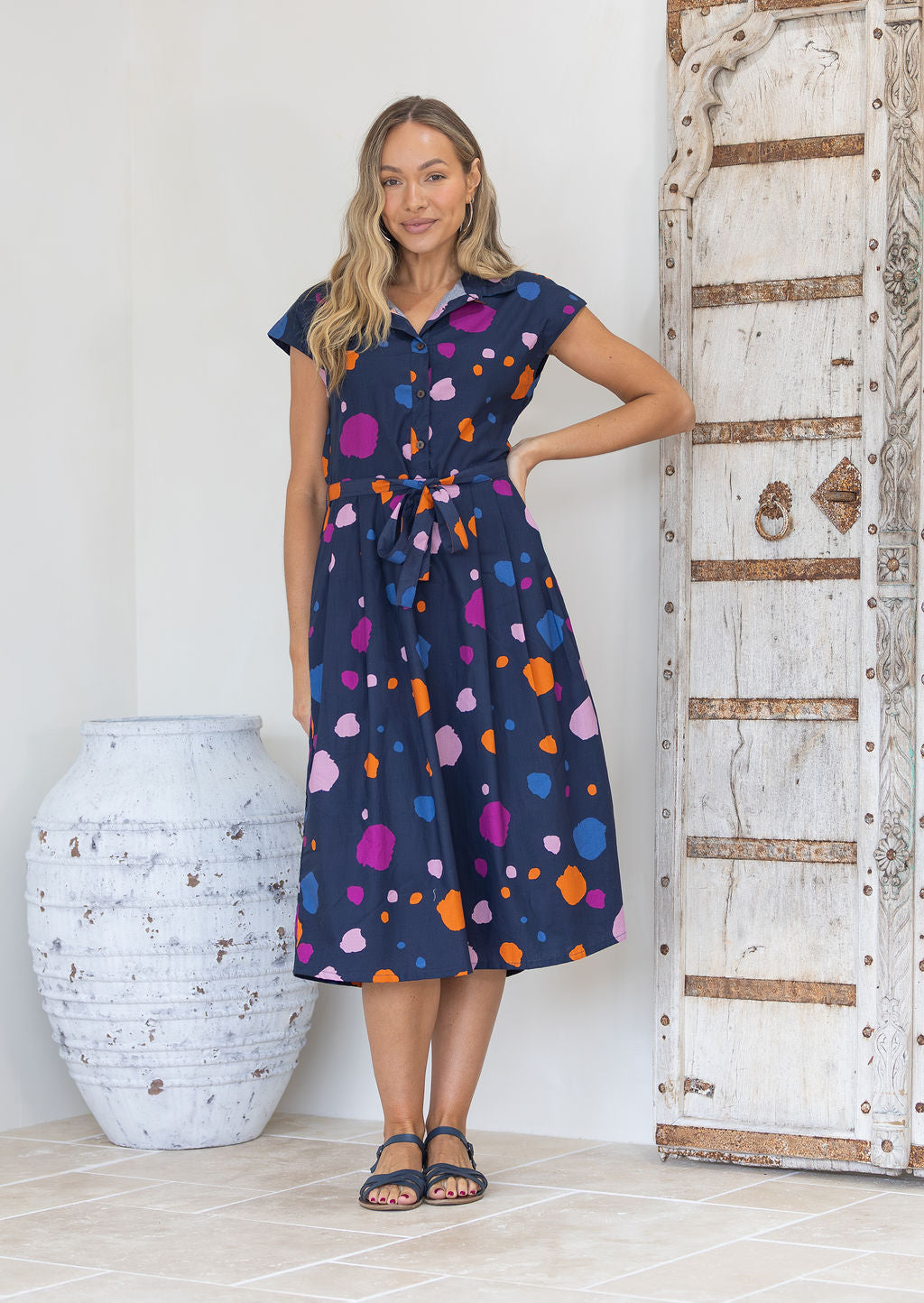 Woman wearing a midi length cotton day dress with a buttoned bodice, cap sleeves, collar and waist tie.