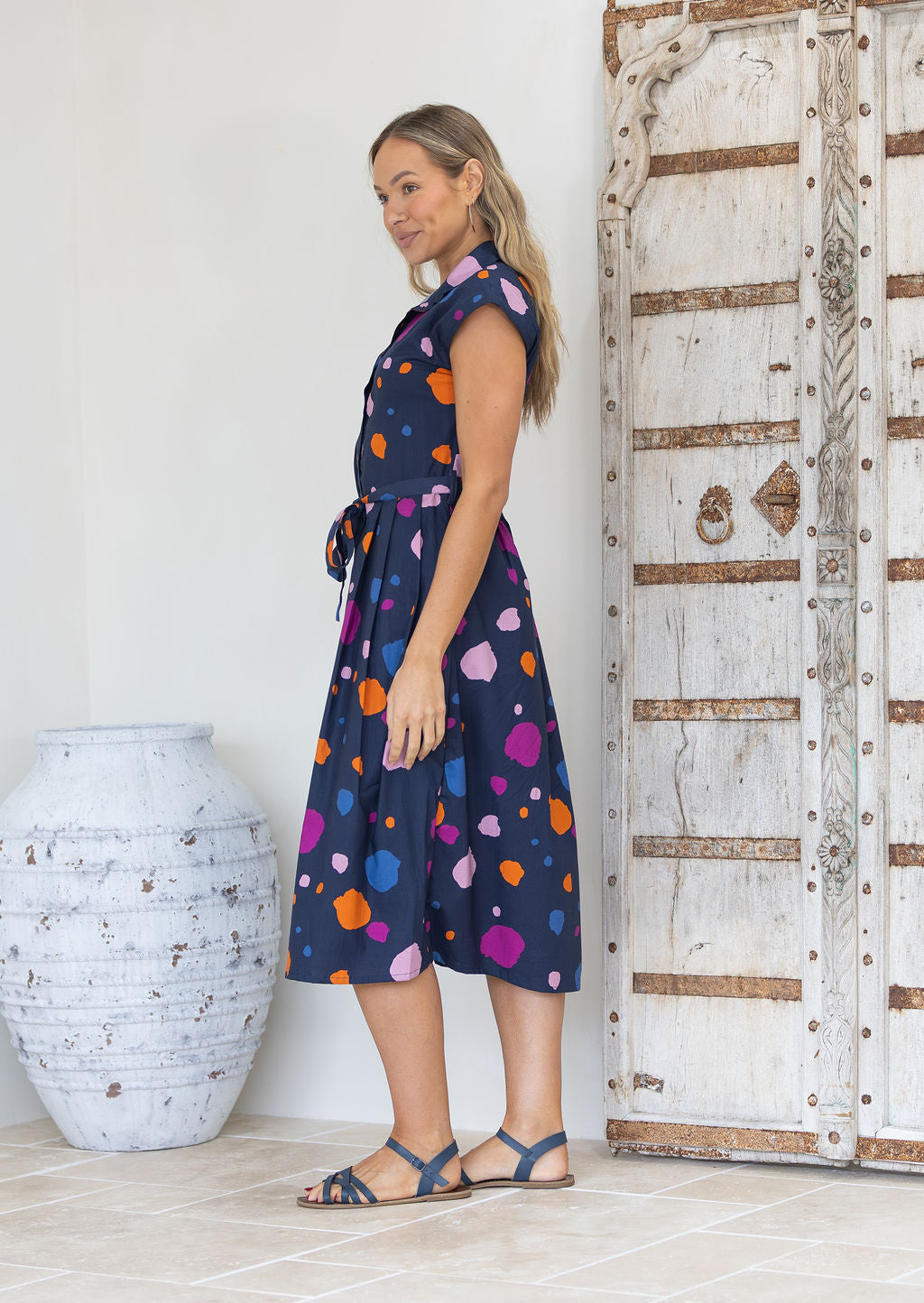 Woman stands side on in a navy based cotton midi length dress with colourful spots