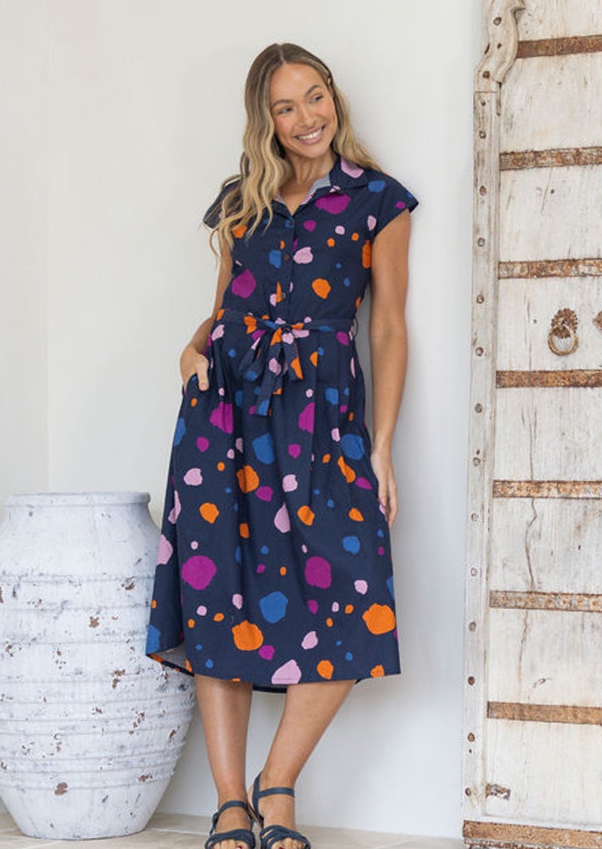 Woman wearing a navy cotton collared dress with colourful polka dots standing against a white wall with hand in pocket