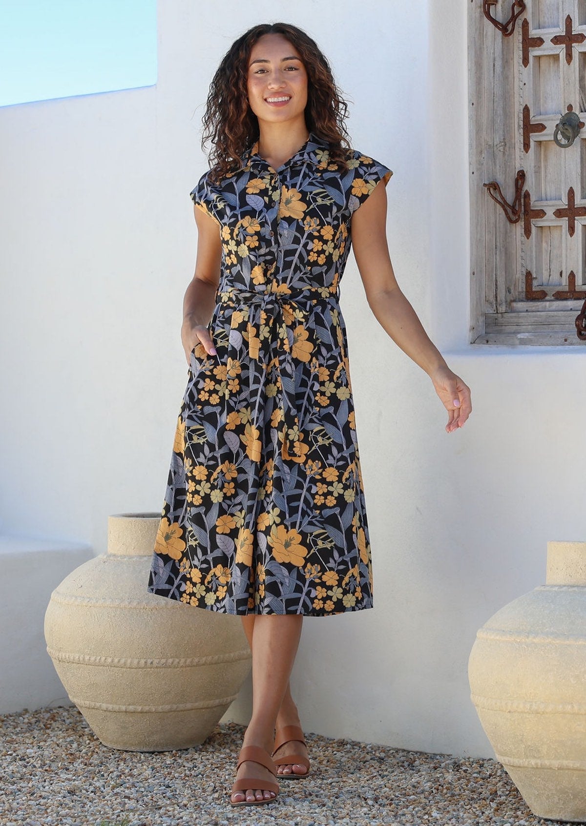 Model stands with one hand in pocket wearing a midi length floral printed shirt style cotton dress with cap sleeves, buttoned bodice and a removable waist tie