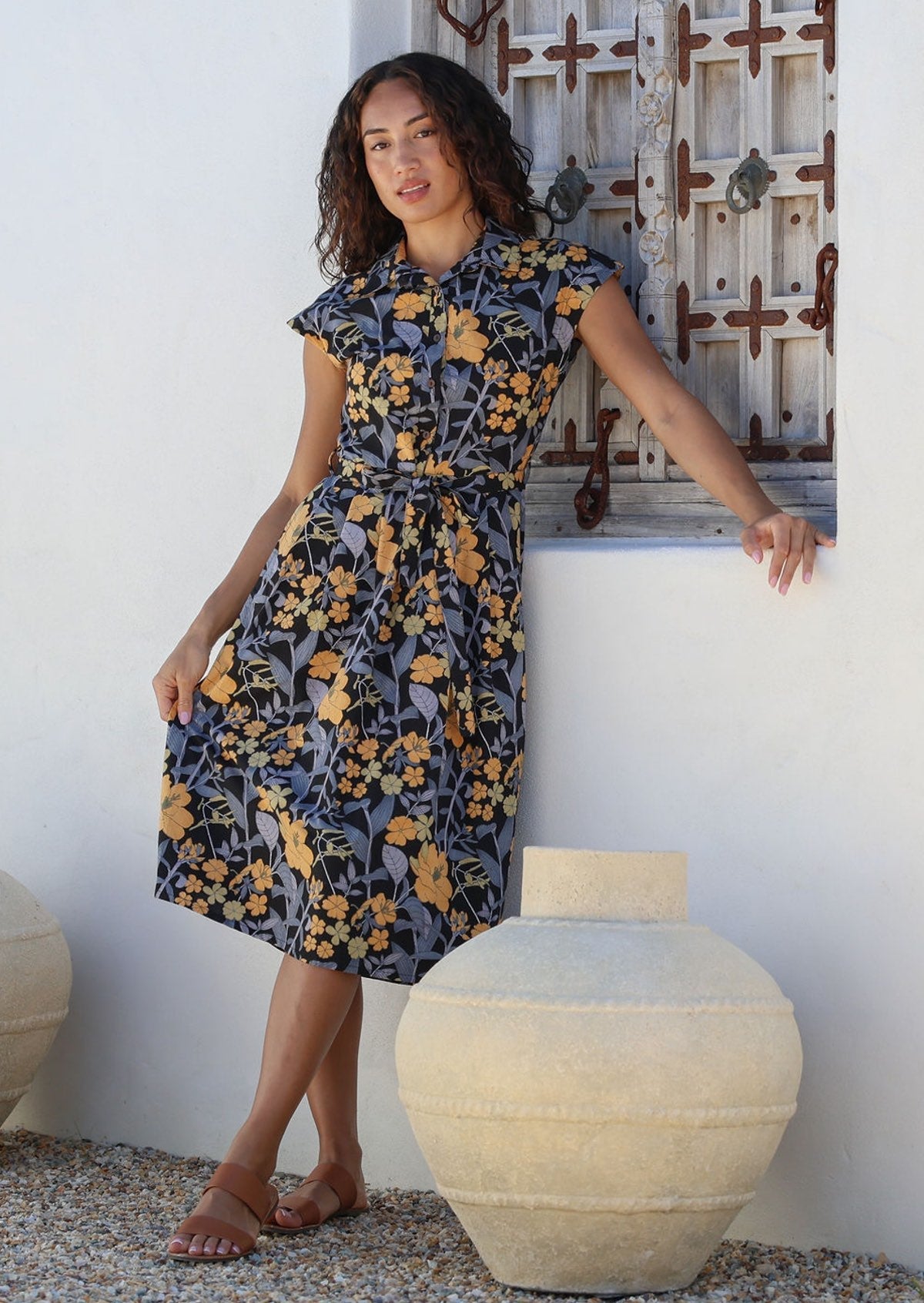 Woman leans on a wall wearing a shirt style cotton midi length dress with buttoned bodice, pockets, collar and cap sleeves