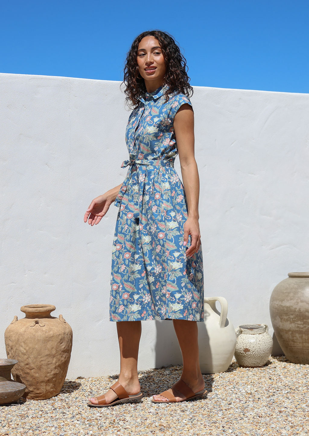 Model stands sideways wearing a floral printed blue shirt style cotton dress with collar, buttoned bodice, pockets and cap sleeves