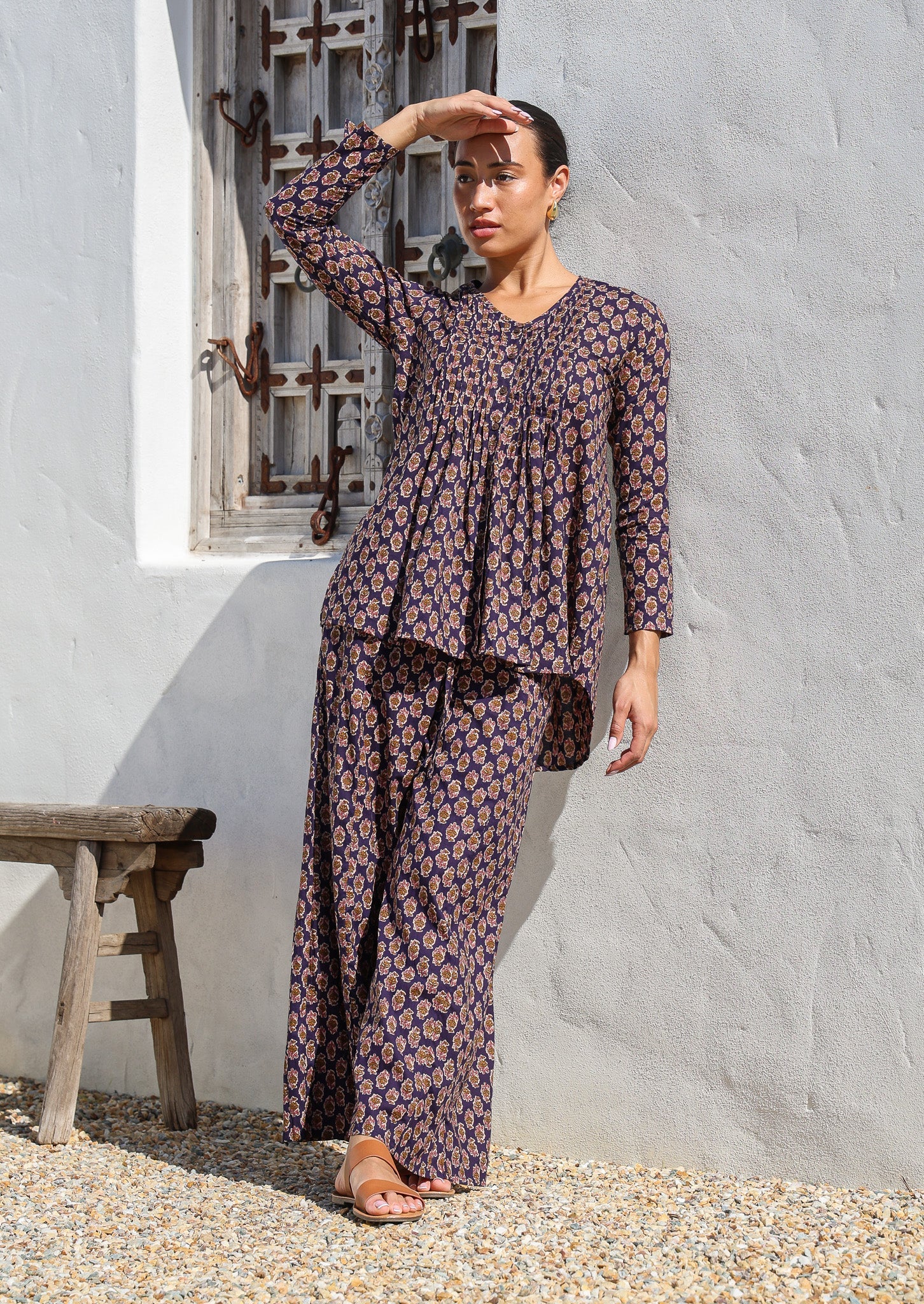 Model standing in sun wears long sleeve button through top with V-neck and small pleats across chest that leads into A line flare at the base.