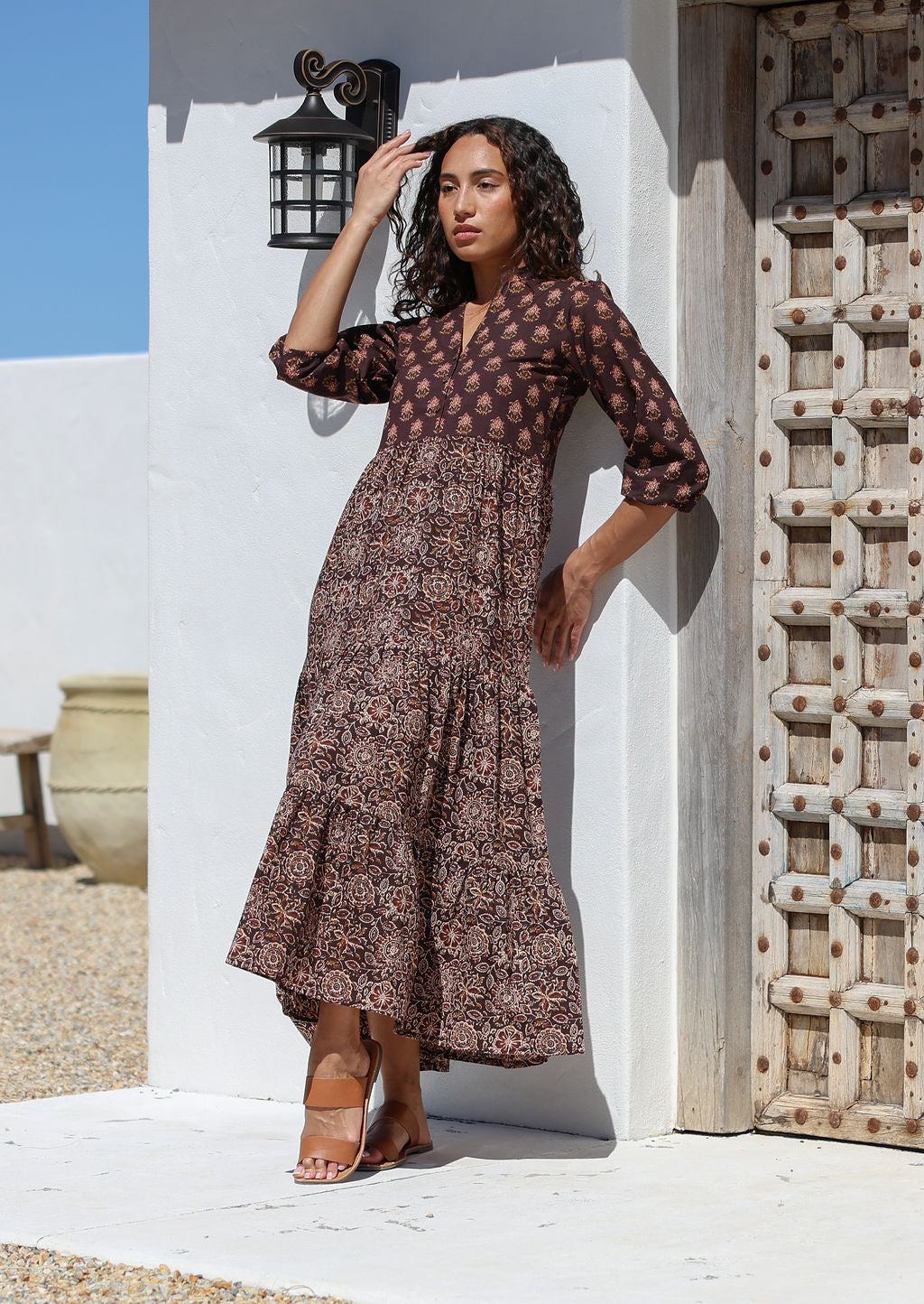Woman in a floral cotton dress standing against a white wall with a wooden door.