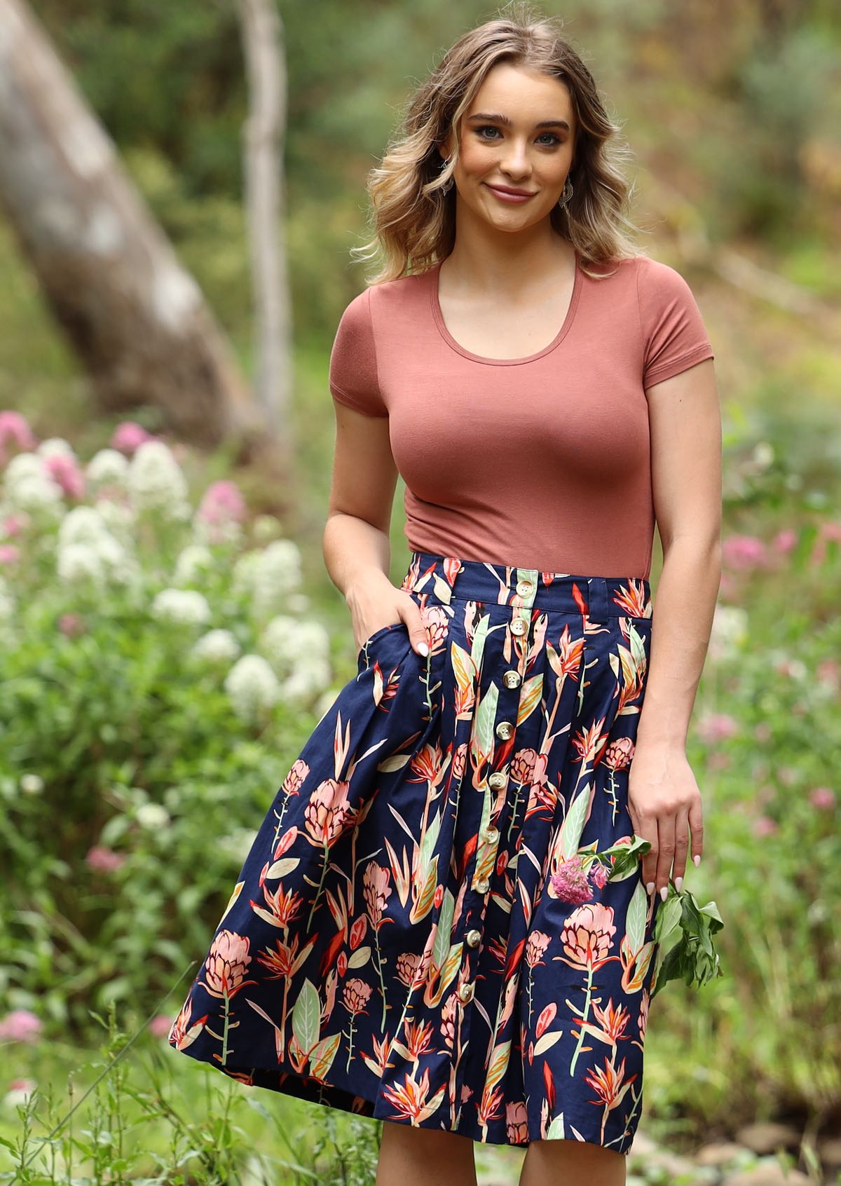 Woman wears knee length button through Ruby skirt with belt loops, box pleats and pockets in botanic print with navy base
