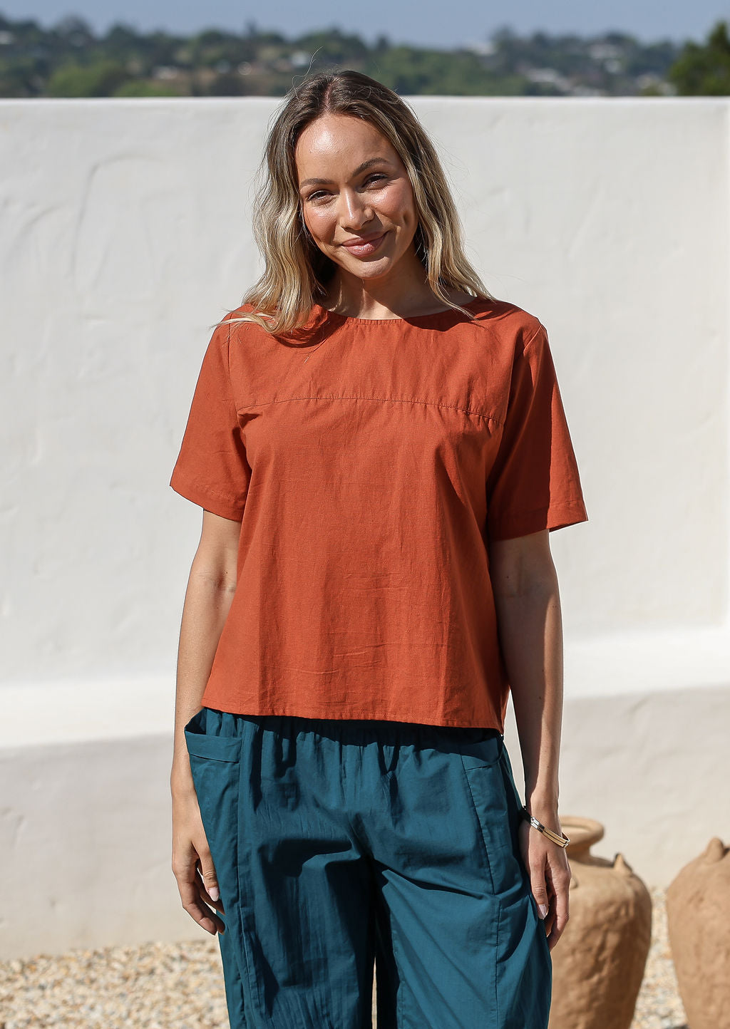 Woman wearing a blue pants and rust shade short sleeve top standing against a white background