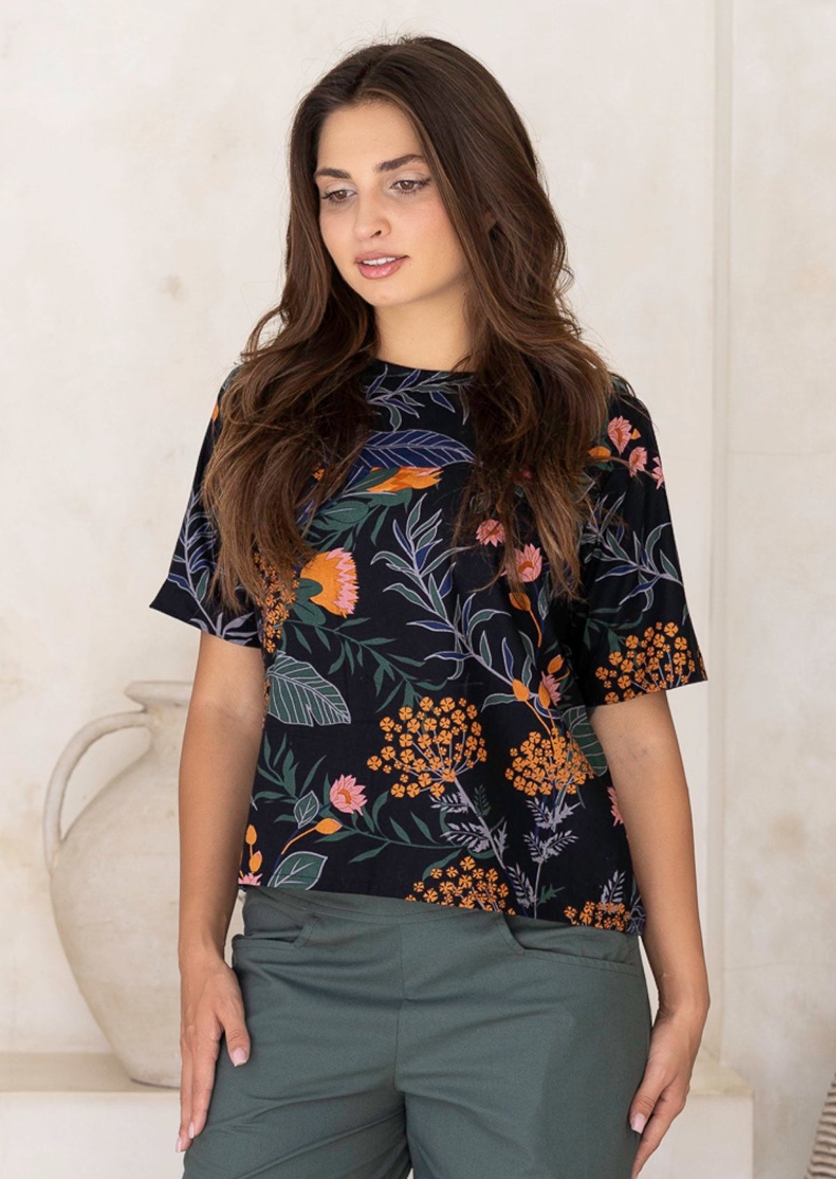 Woman wearing a loose fit black floral with green pants against a white wall.