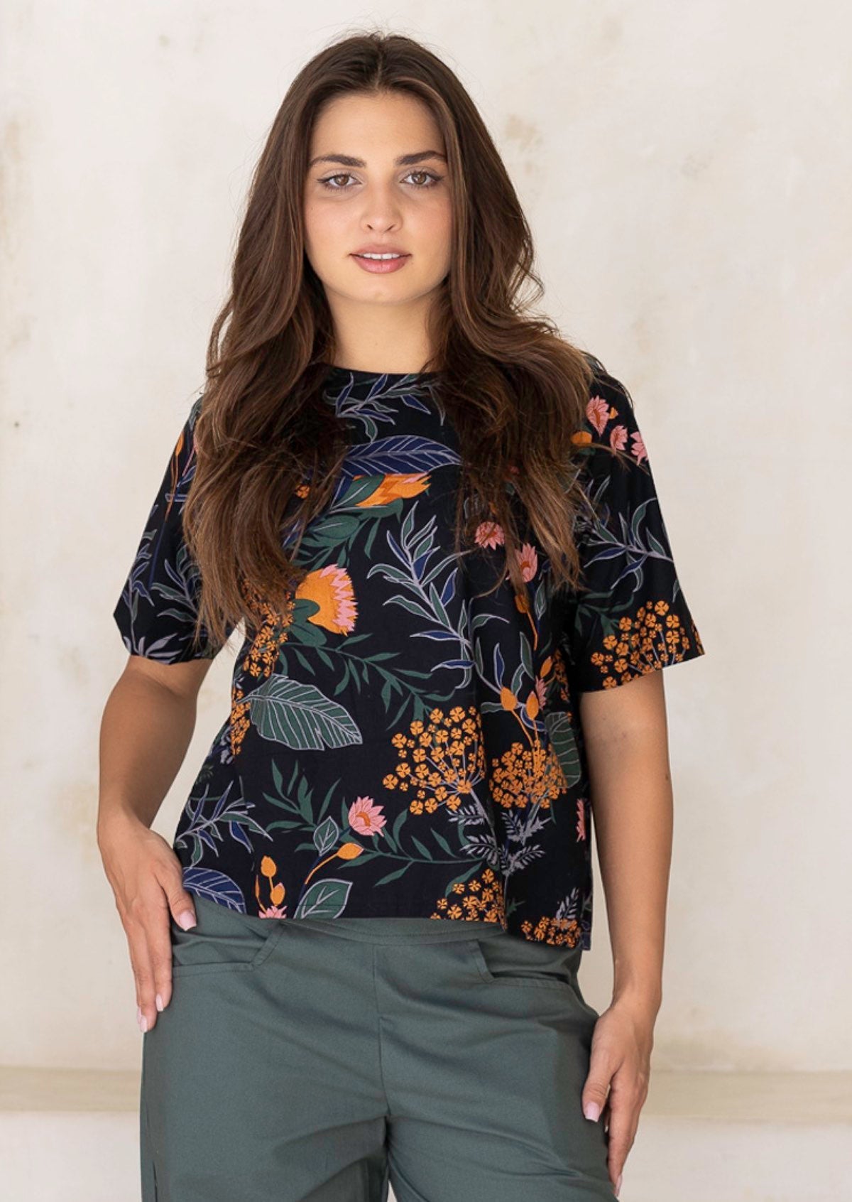 Woman posing in a 100 percent cotton loose cut blouse with beautiful botanical print in blue, green, greys with orange and pink florals on black base.
