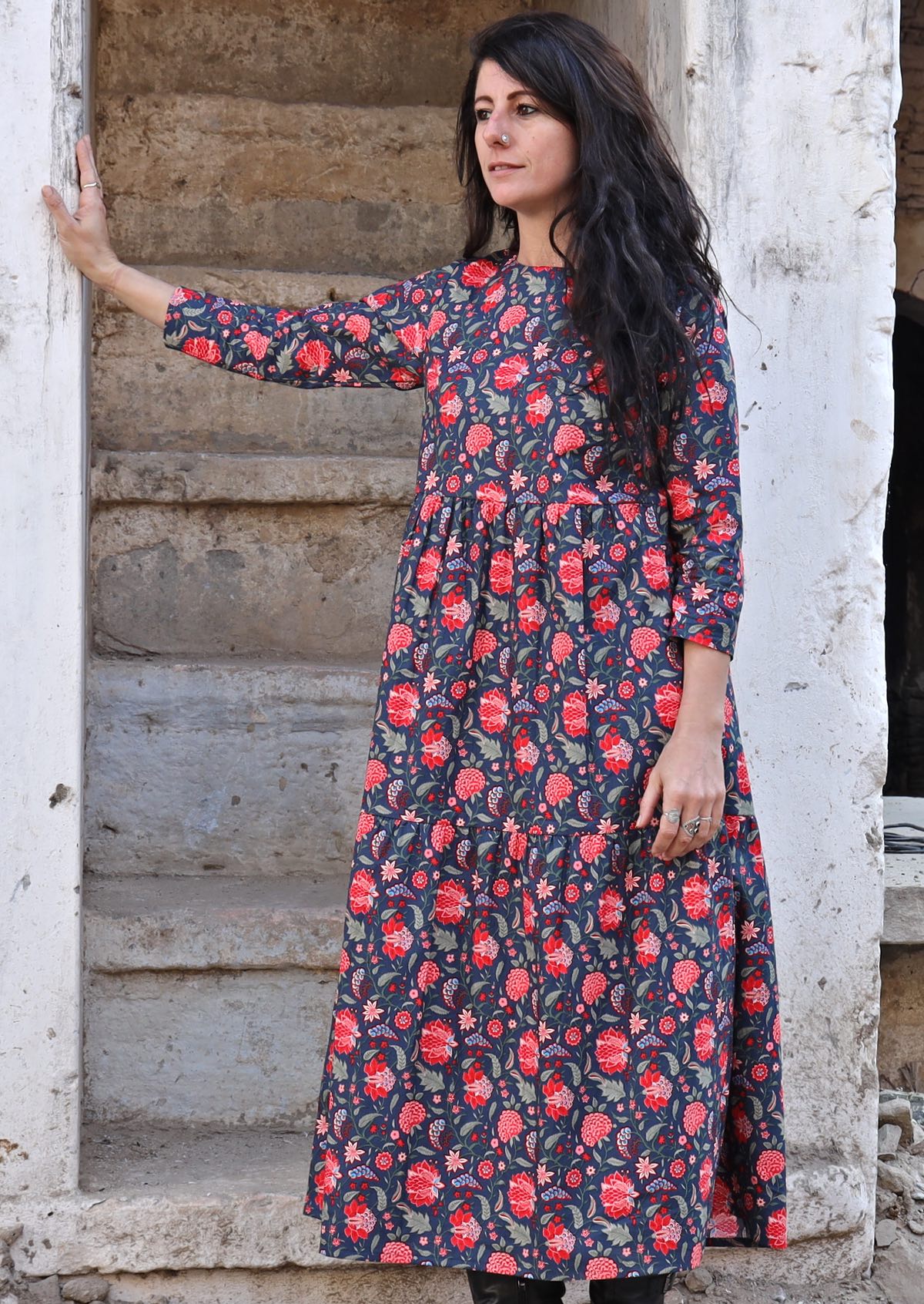 Model standing in front of cement stairs with dark hair wearing a 100 percent cotton three tiered midi length dress with blue base and red floral print
