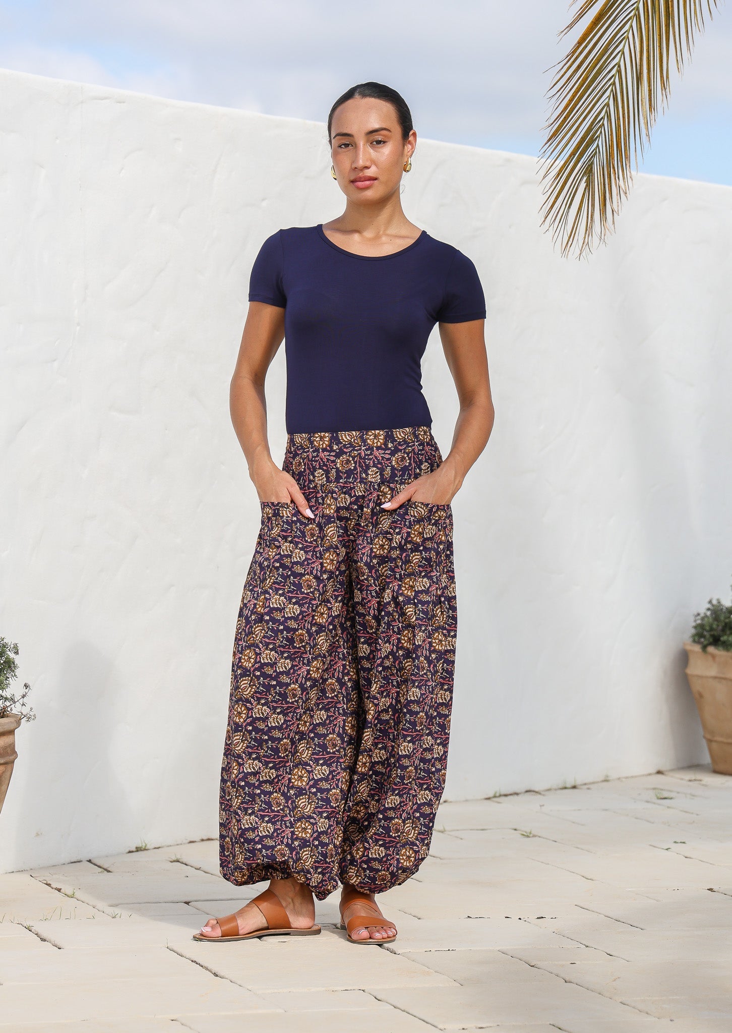 Model standing in front of white wall wears harem style cotton pants in deep purple based floral print with hands in pockets.