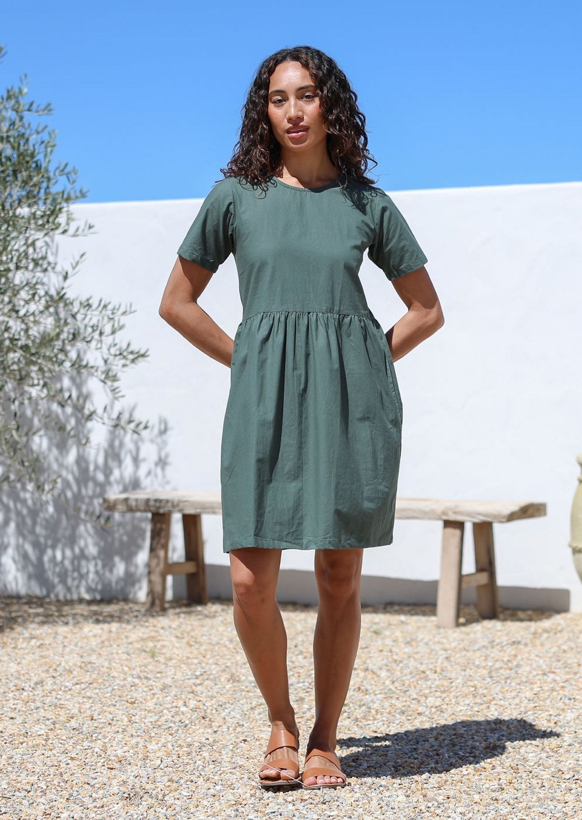 Model with her hands at the back wears short sleeved green cotton dress with round neckline and small gathers at the waist