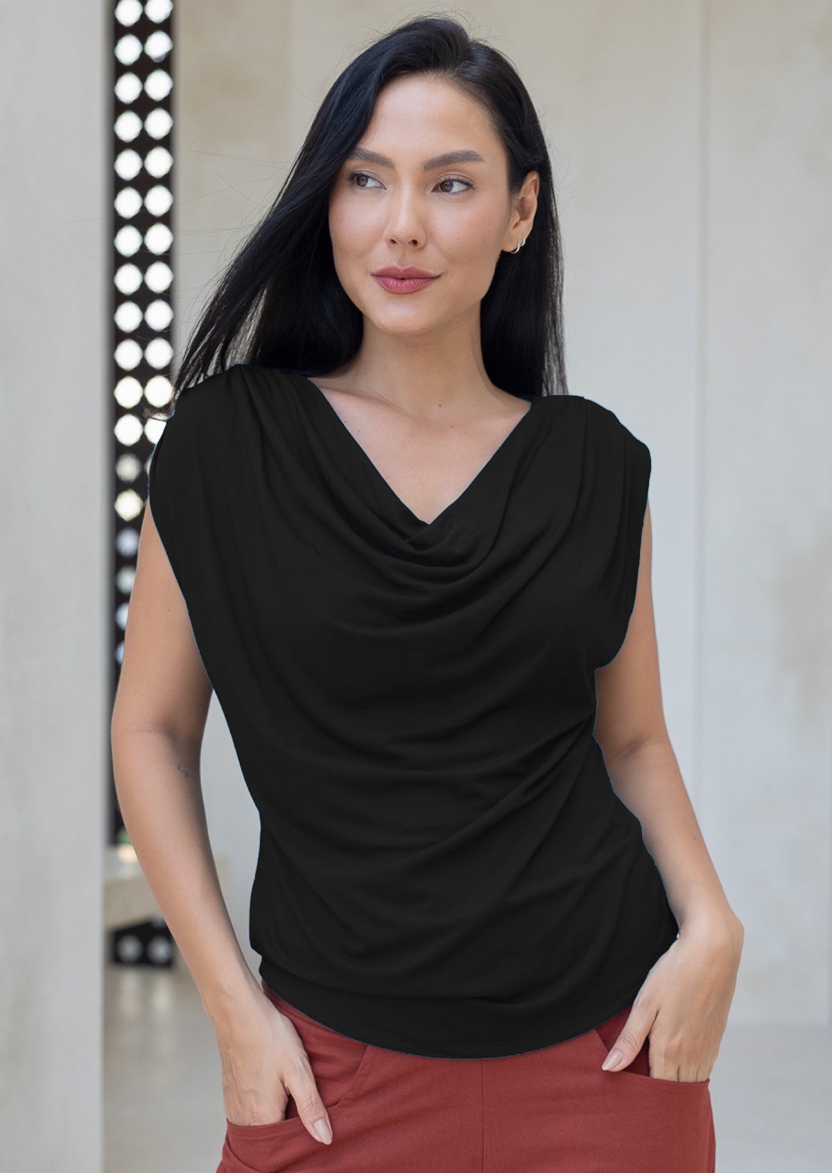 Woman wearing a black sleeveless top with a draped neckline, with hands in pockets of rust pants