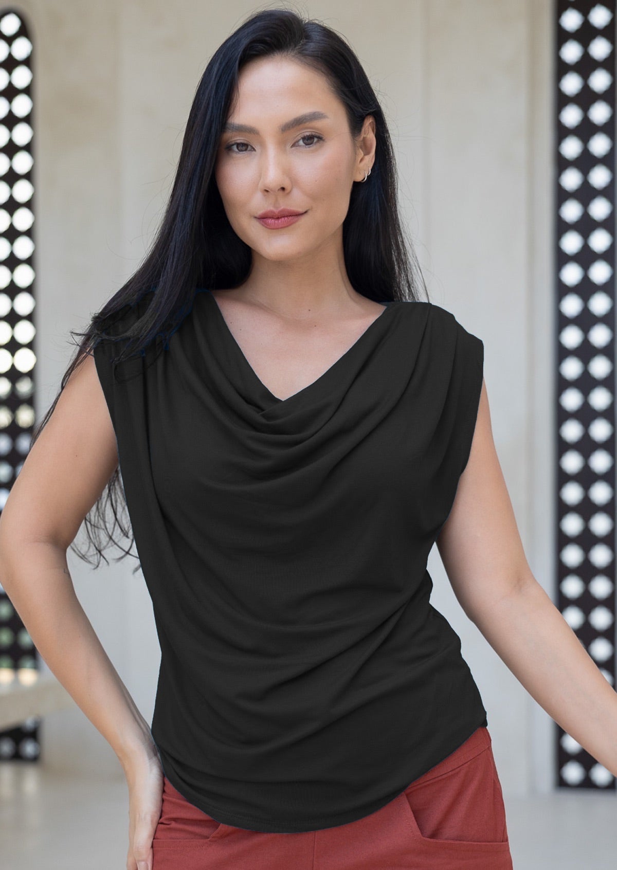 Woman wearing a black sleeveless top with a draped neckline, standing against a patterned wall.