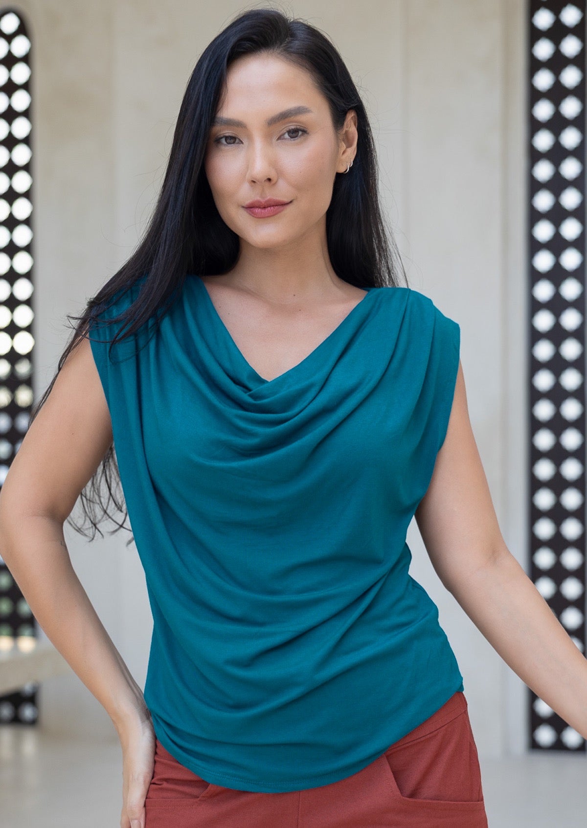 Woman wearing a teal blue sleeveless top with a draped neckline, standing against a neutral background.