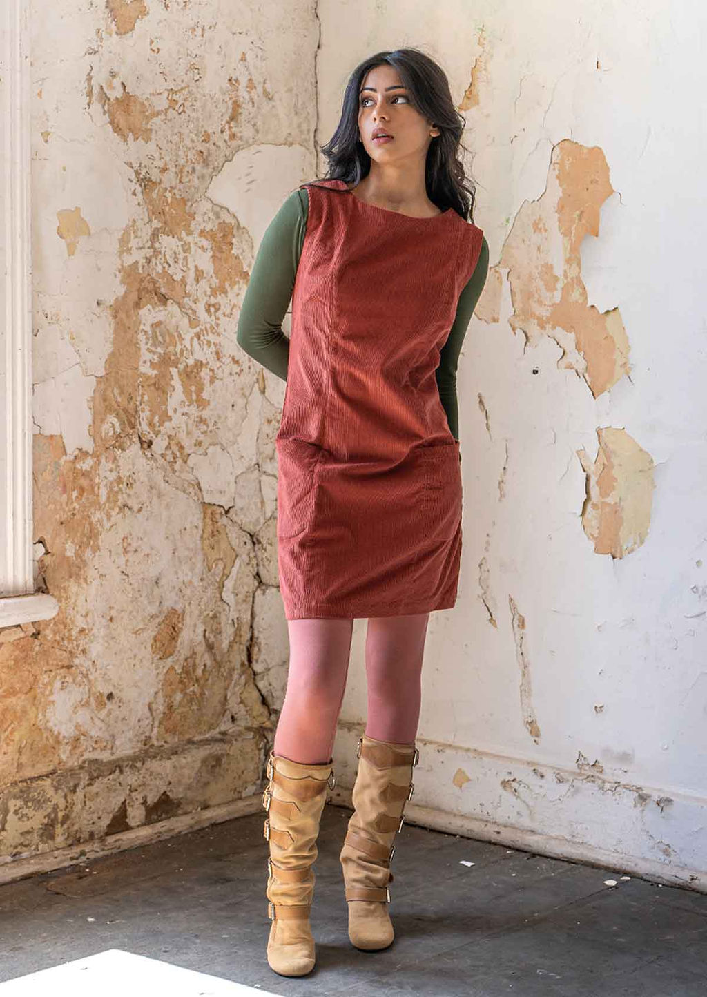 Model wears burnt henna corduroy sleeveless tunic with olive long sleeve top and dusty pink legging and boots