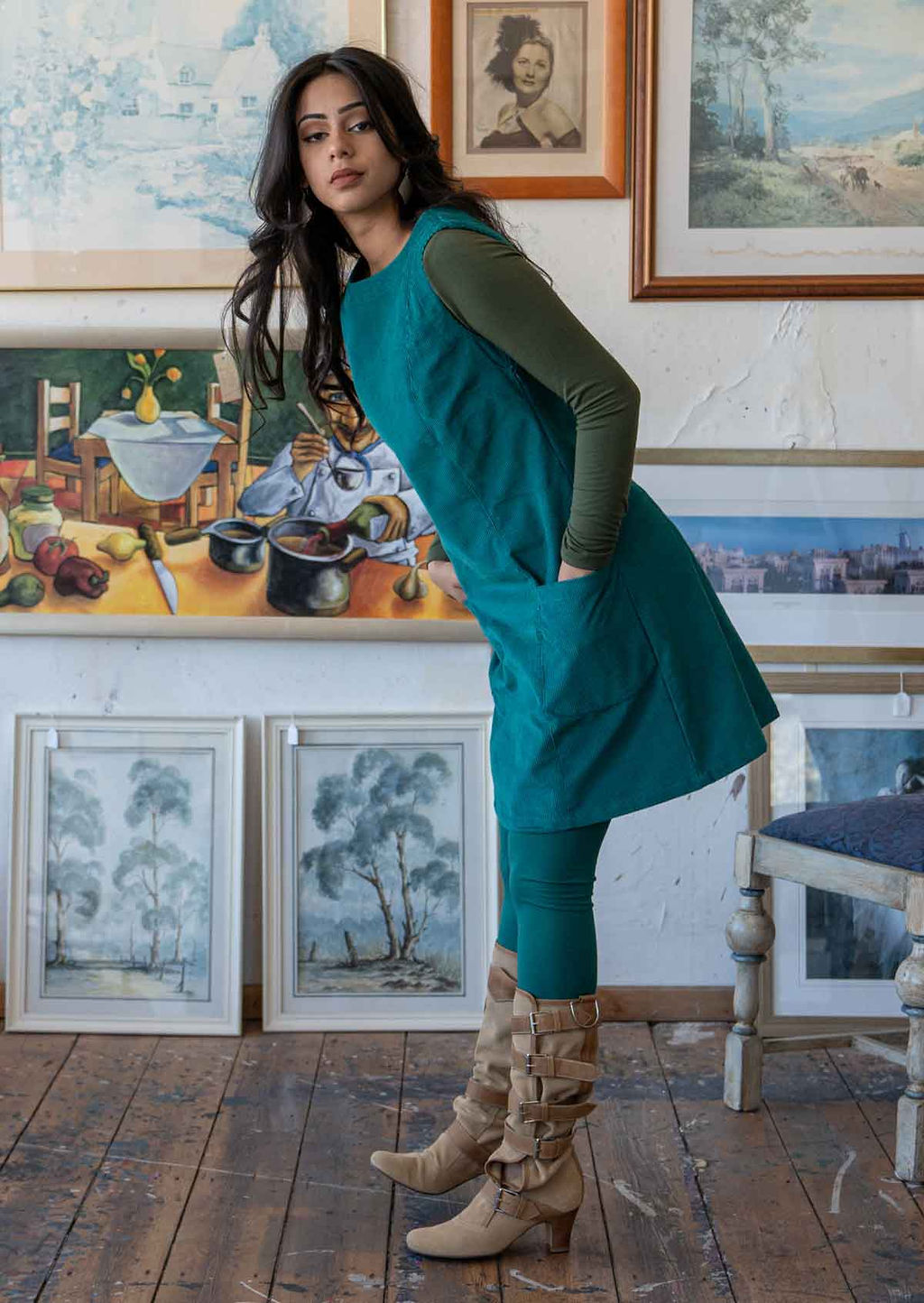 Woman wears corduroy sleeveless tunic with high round neckline and pockets in teal colour with olive long sleeve top underneath, leggings and boots