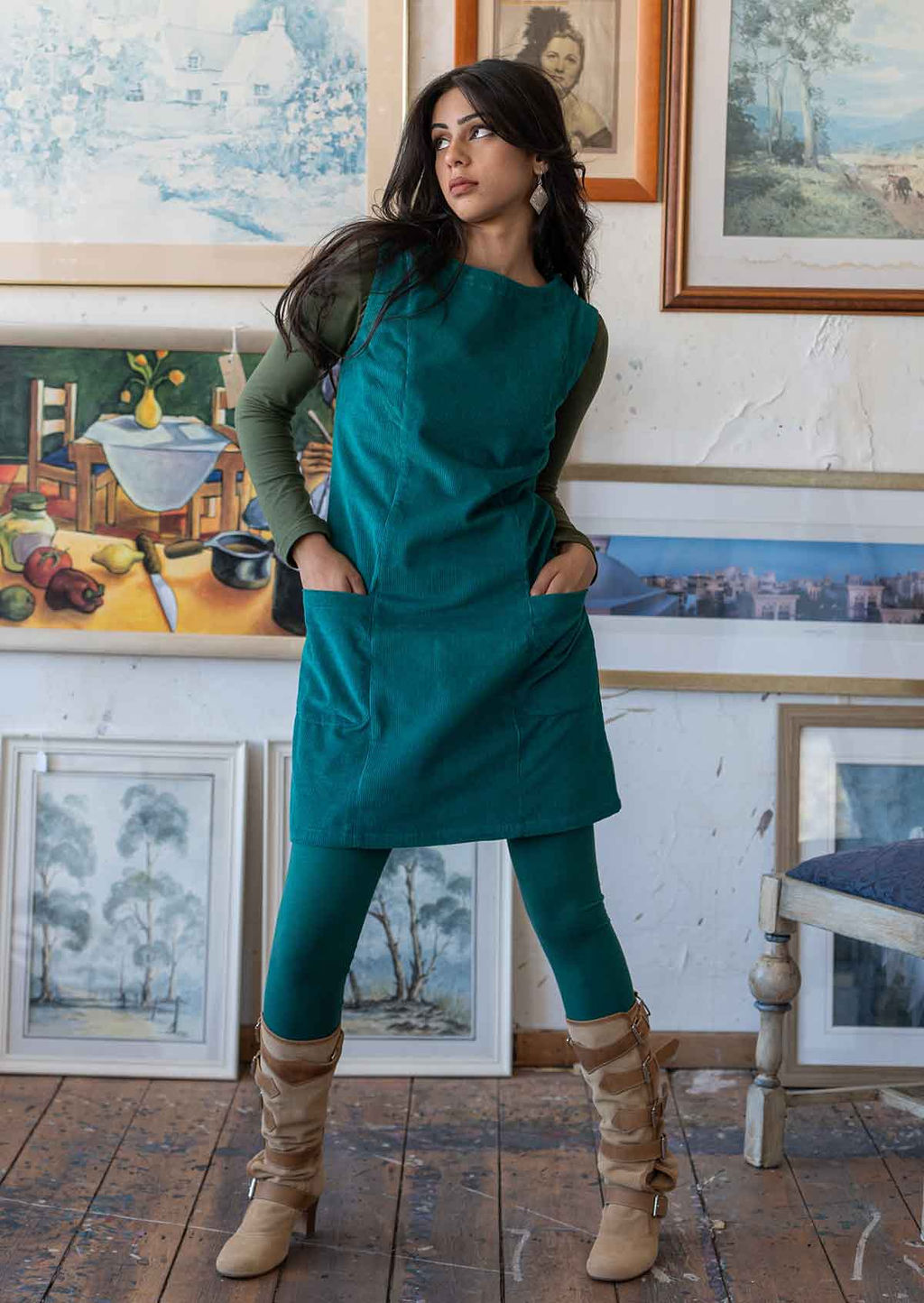 Model poses in sleeveless corduroy tunic with hands in pockets, paired with an olive long sleeve top underneath and leggings and boots
