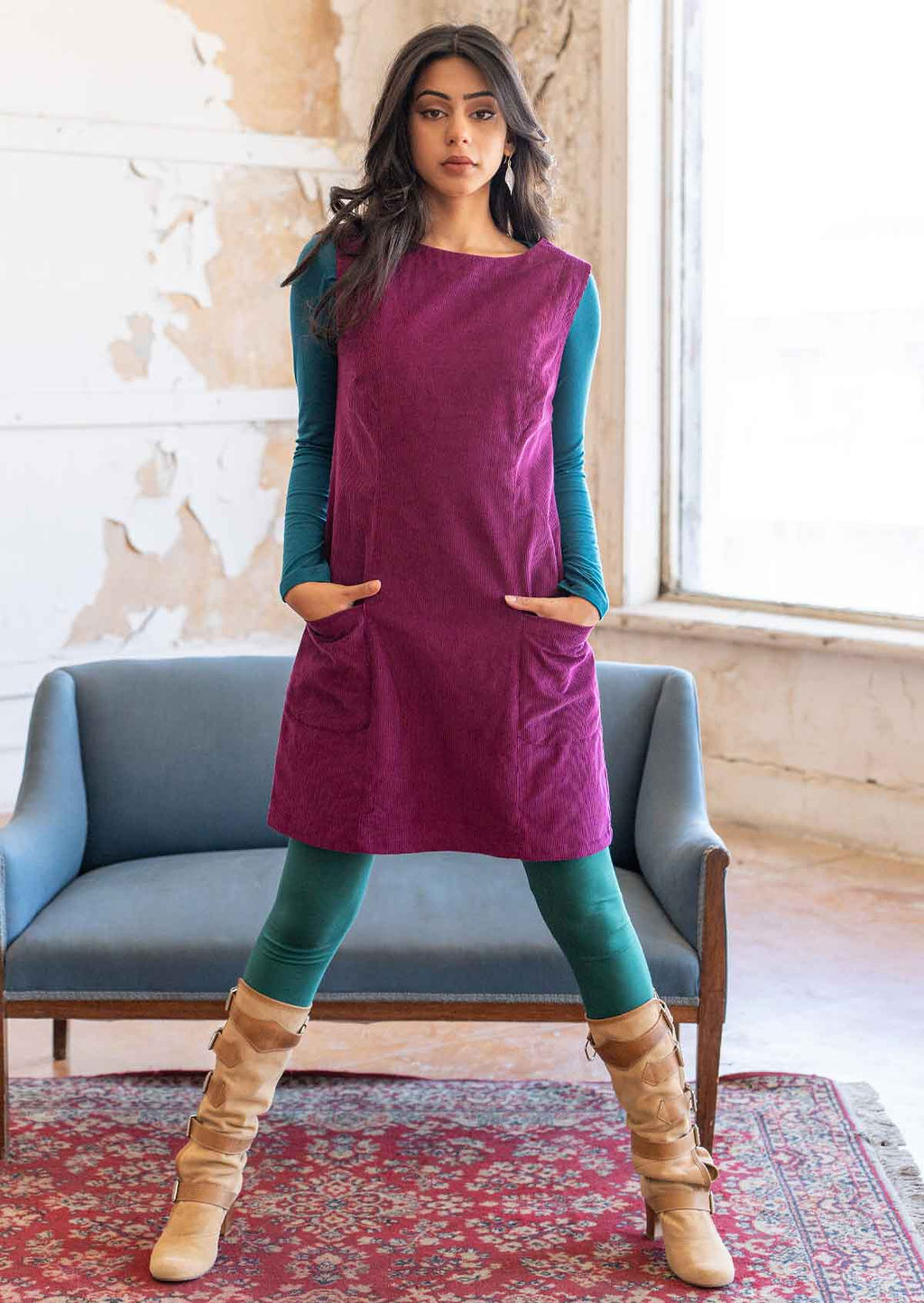 Woman poses in plum purple corduroy sleeveless tunic with hands in pockets, paired with long sleeve teal top underneath, jade leggings and boots