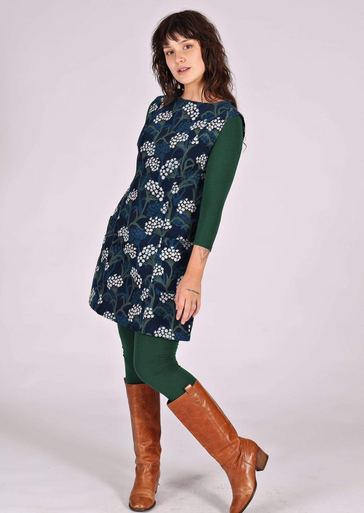 Woman stands in floral print cotton corduroy sleeveless tunic layered over dark green leggings and long sleeve top