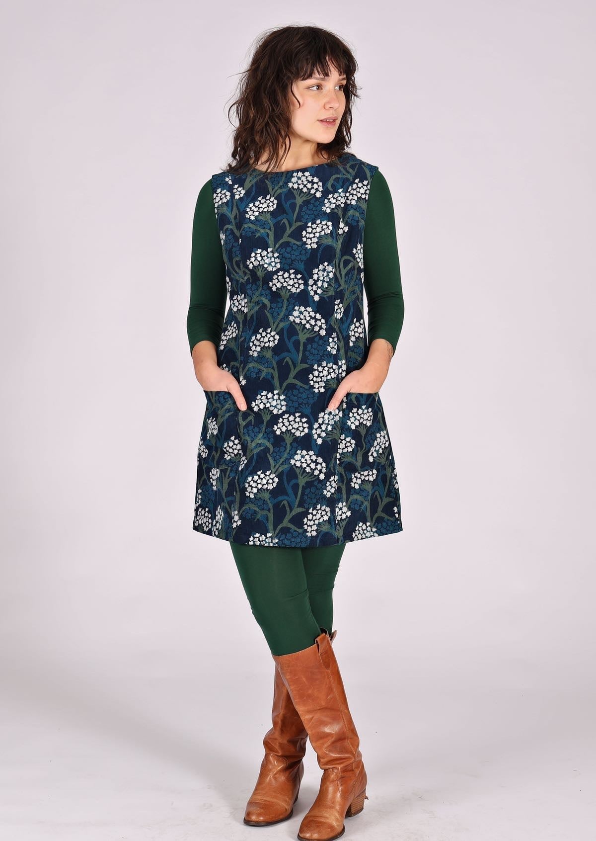 Woman stands in cotton corduroy sleeveless tunic layered over dark green leggings and long sleeve top. with hands in pockets