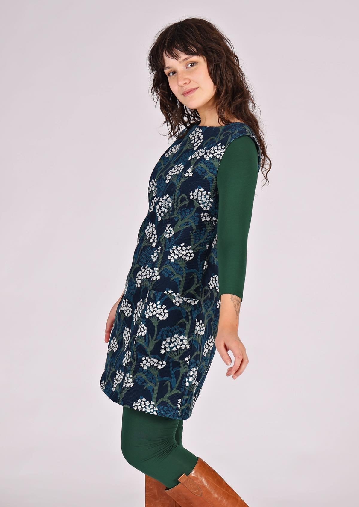 Woman wears floral print cotton corduroy sleeveless tunic layered over dark green leggings and long sleeve top