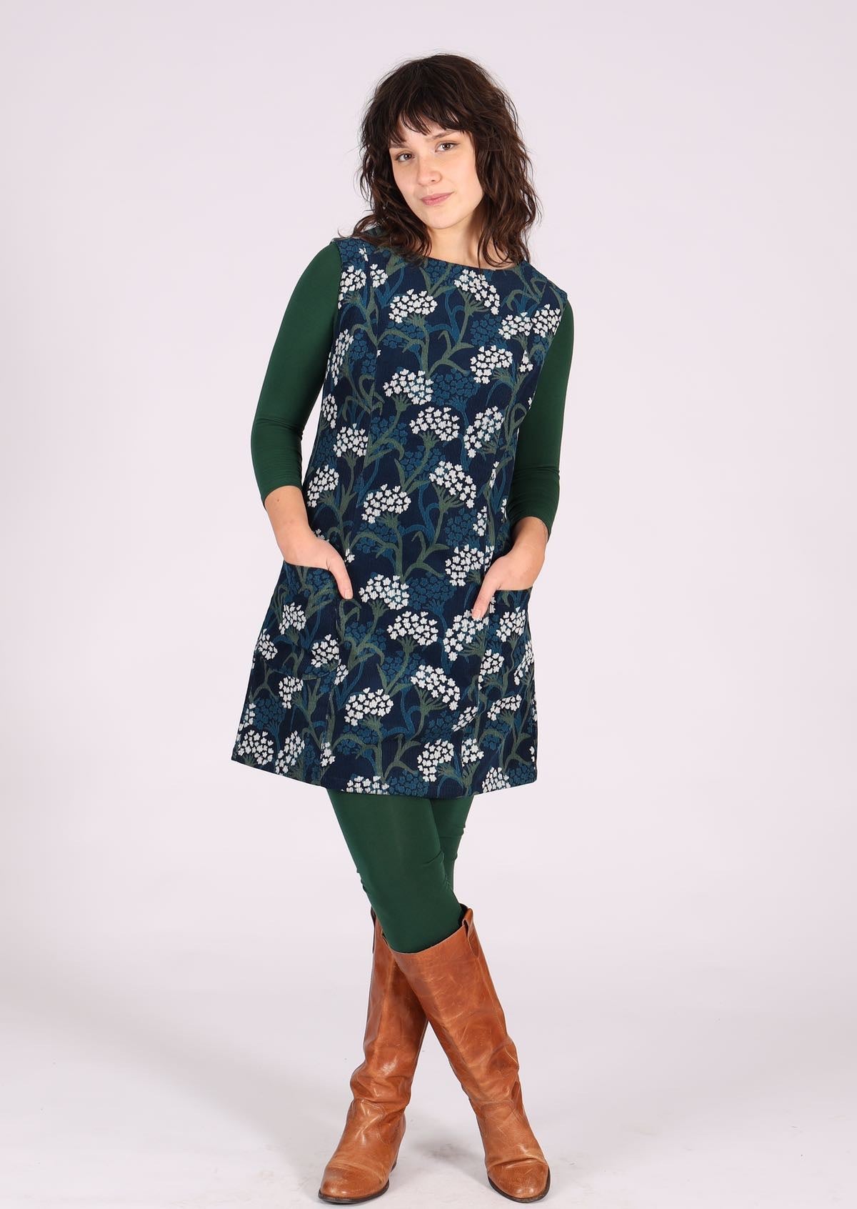 Woman poses in floral print cotton corduroy sleeveless tunic layered with dark green leggings, long sleeve top and boots