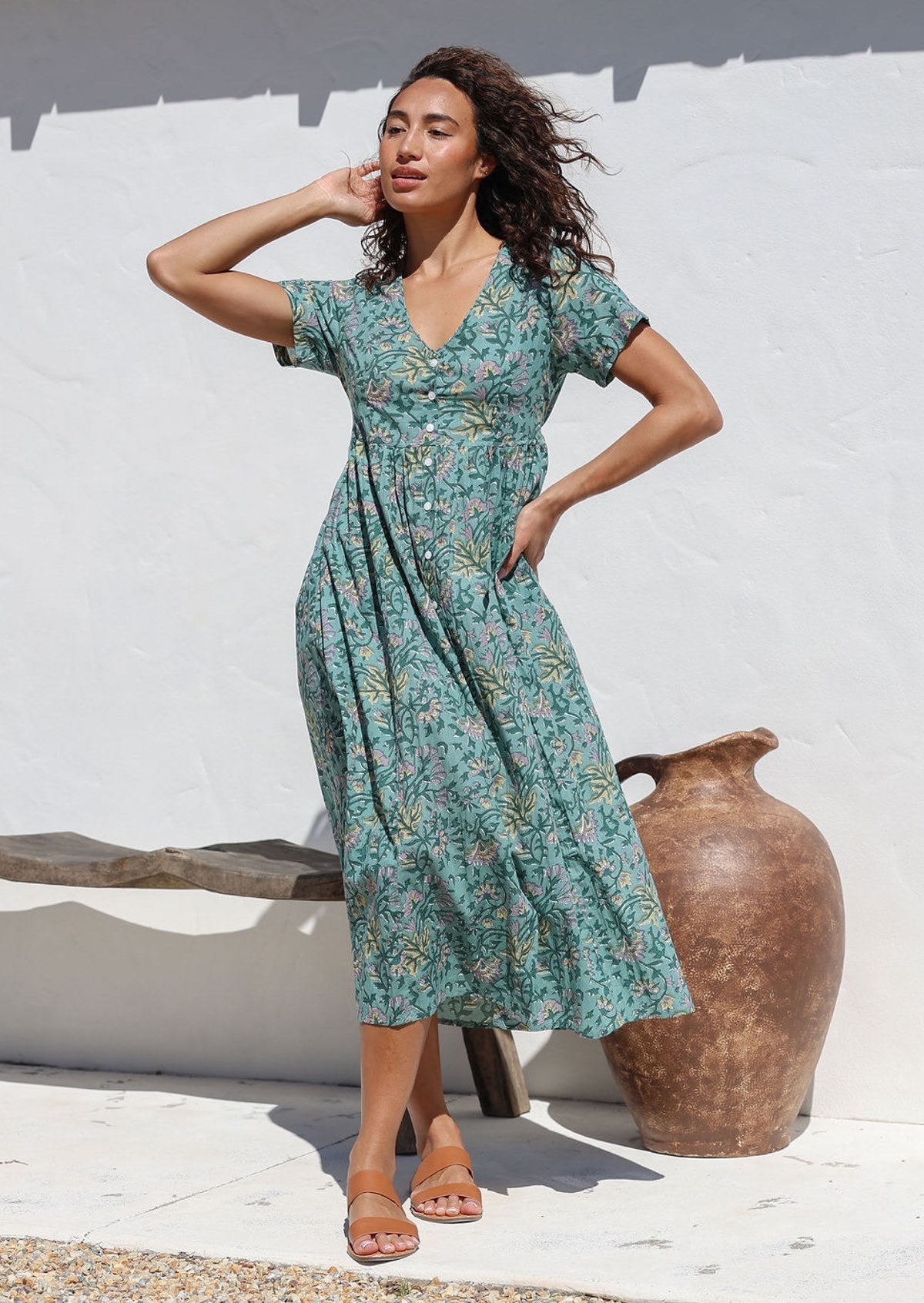 Woman wearing floral cotton midlength dress with short sleeves, shoestring tie and button front standing in front of white wall and wooden bench