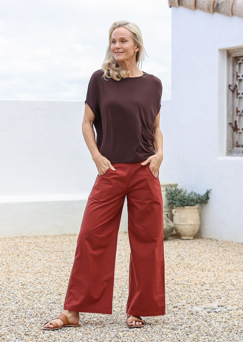 Woman standing with hands in pockets  wearing a brown top and wide legged cotton remi pants in burnt henna shade with side zipper.