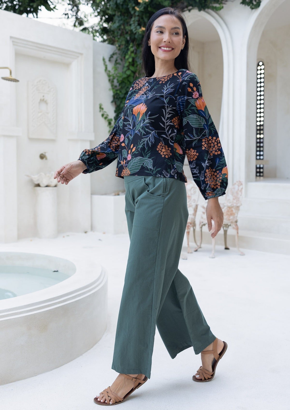 Woman walking in a black based floral top and green wide legged pants in a white courtyard.
