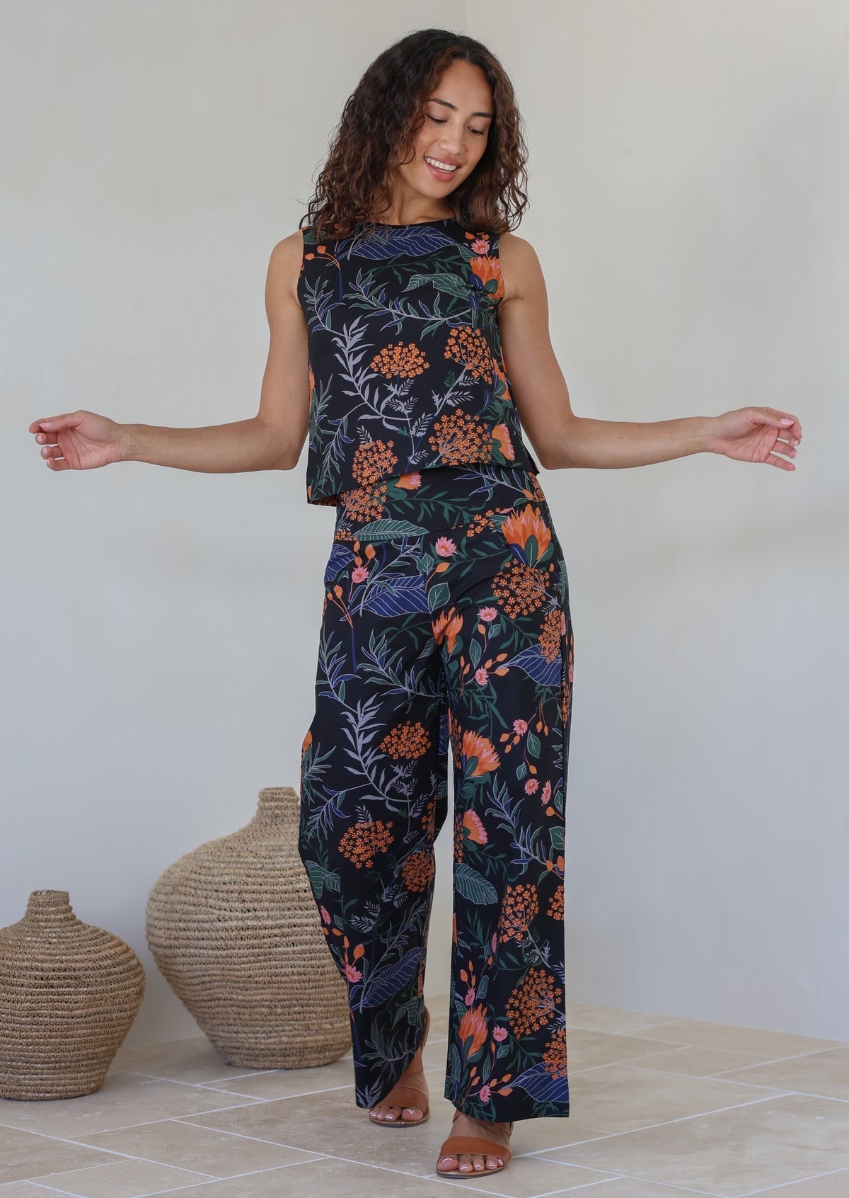 The model wears a matching botanical print top and high waisted wide leg cotton pants in black with orange, pink, green, and blue prints