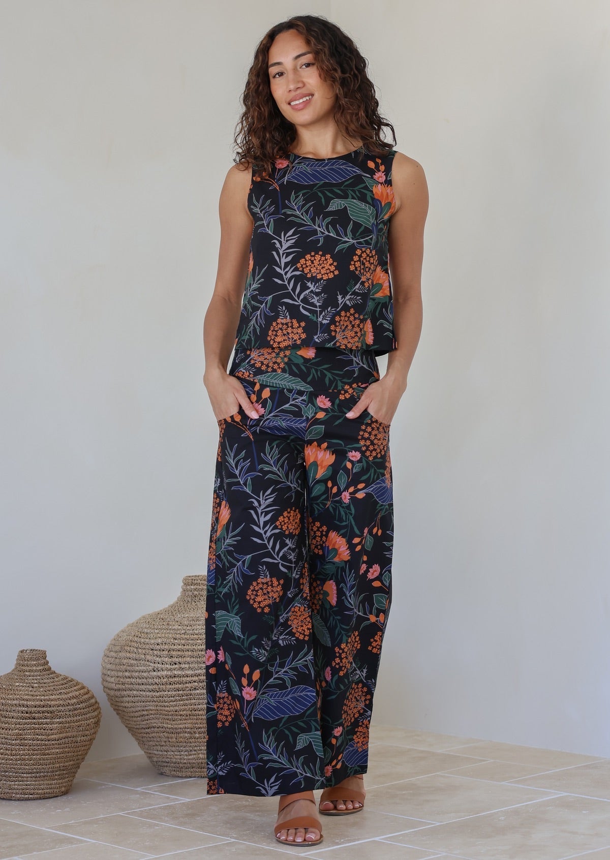 Model stands with both the hands in pocket wearing a 100% cotton wide leg pants in black based colorful botanical print
